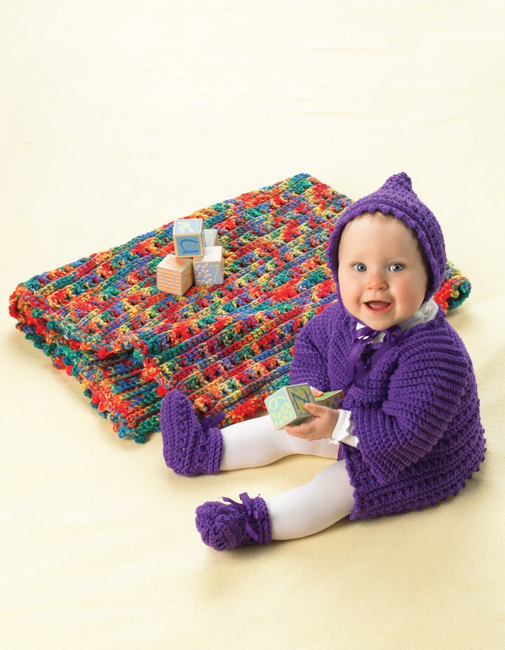 A smiling baby in a purple outfit sits on a pale yellow surface, holding a block. Beside them is a colorful blanket crocheted from "Bright Layettes to Crochet" by Leisure Arts, along with a few wooden blocks.