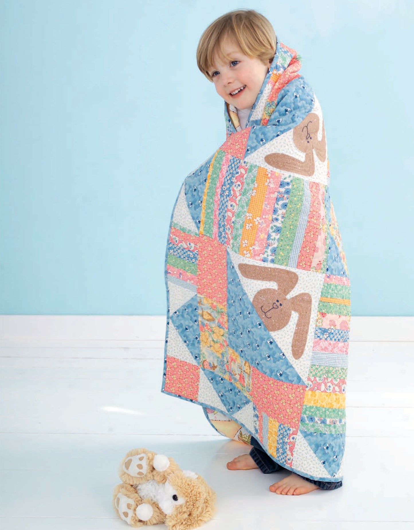 A young child smiles, wrapped in a colorful bunny patchwork from Leisure Arts’ Wee Wonder Quilts Digital Download for infants and toddlers. The scene is set in a light blue room, with a plush toy resting on the white floor.