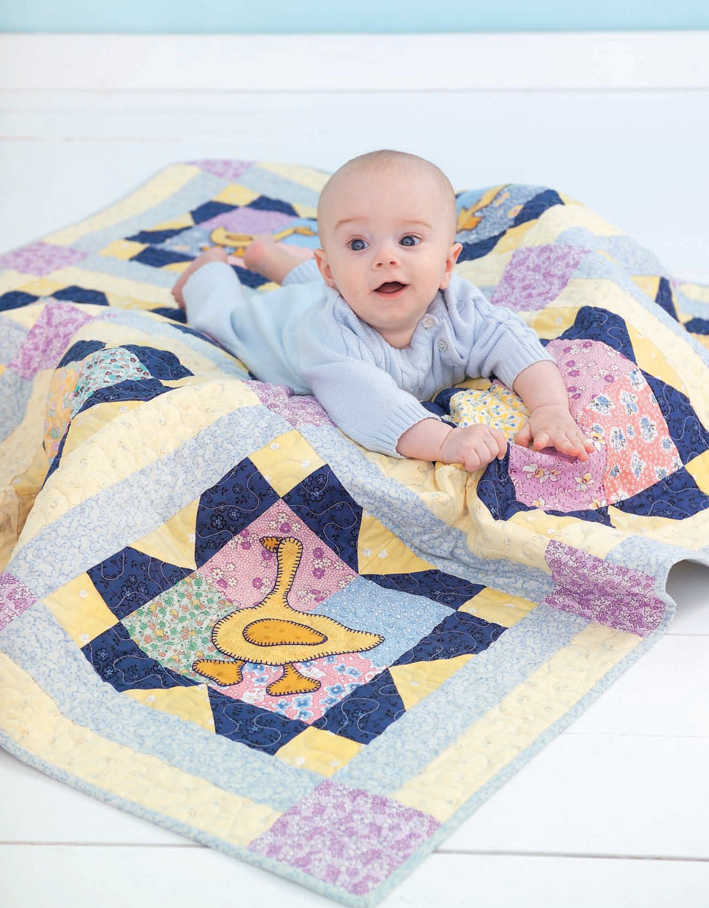 A baby in a light blue outfit lies on a pastel patchwork quilt with playful designs, including a quilted duck. Perfect for infants and toddlers, the Wee Wonder Quilts Digital Download by Leisure Arts offers charming options for little ones.