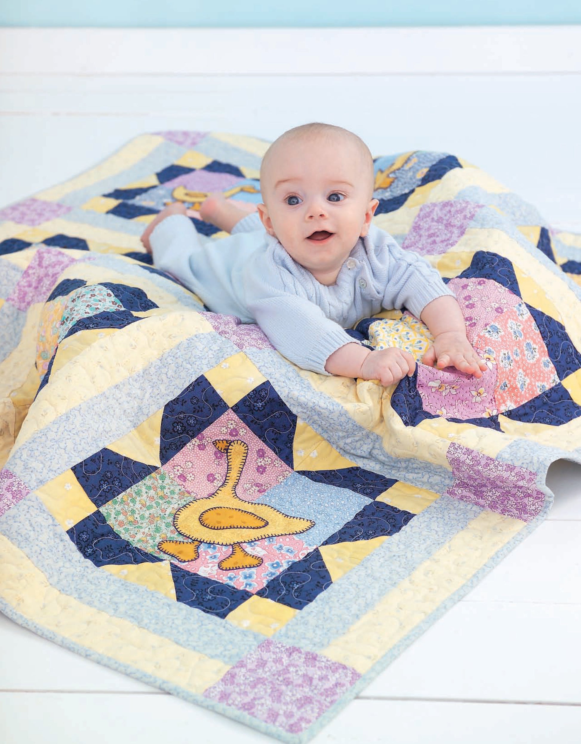 A baby in a light blue outfit lies on a pastel patchwork quilt with playful designs, including a quilted duck. Perfect for infants and toddlers, the Wee Wonder Quilts Digital Download by Leisure Arts offers charming options for little ones.