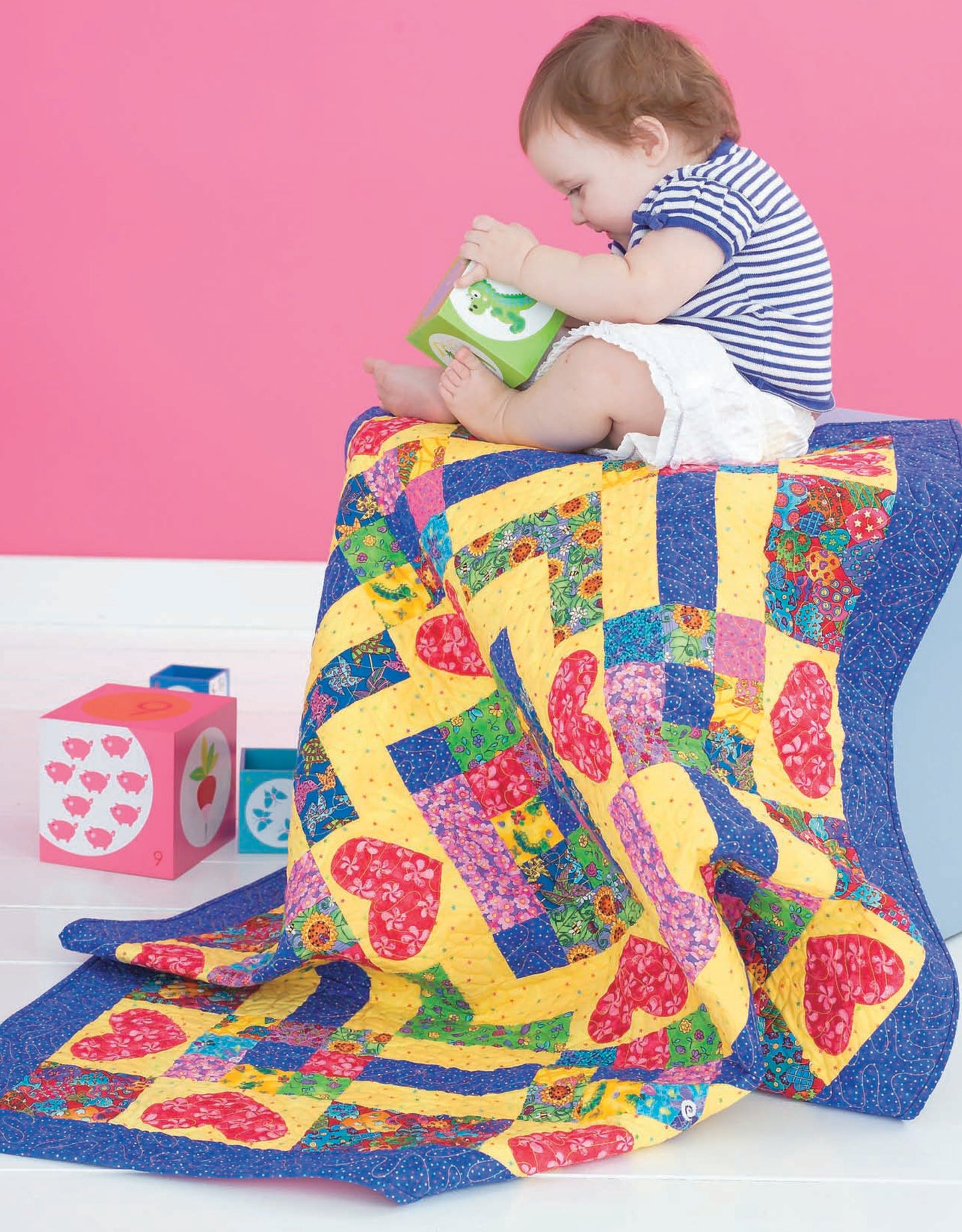 A baby in a striped shirt sits on a chair draped with the Leisure Arts Wee Wonder Quilts Digital Download, playing with bright blocks, all set against a vibrant pink wall.
