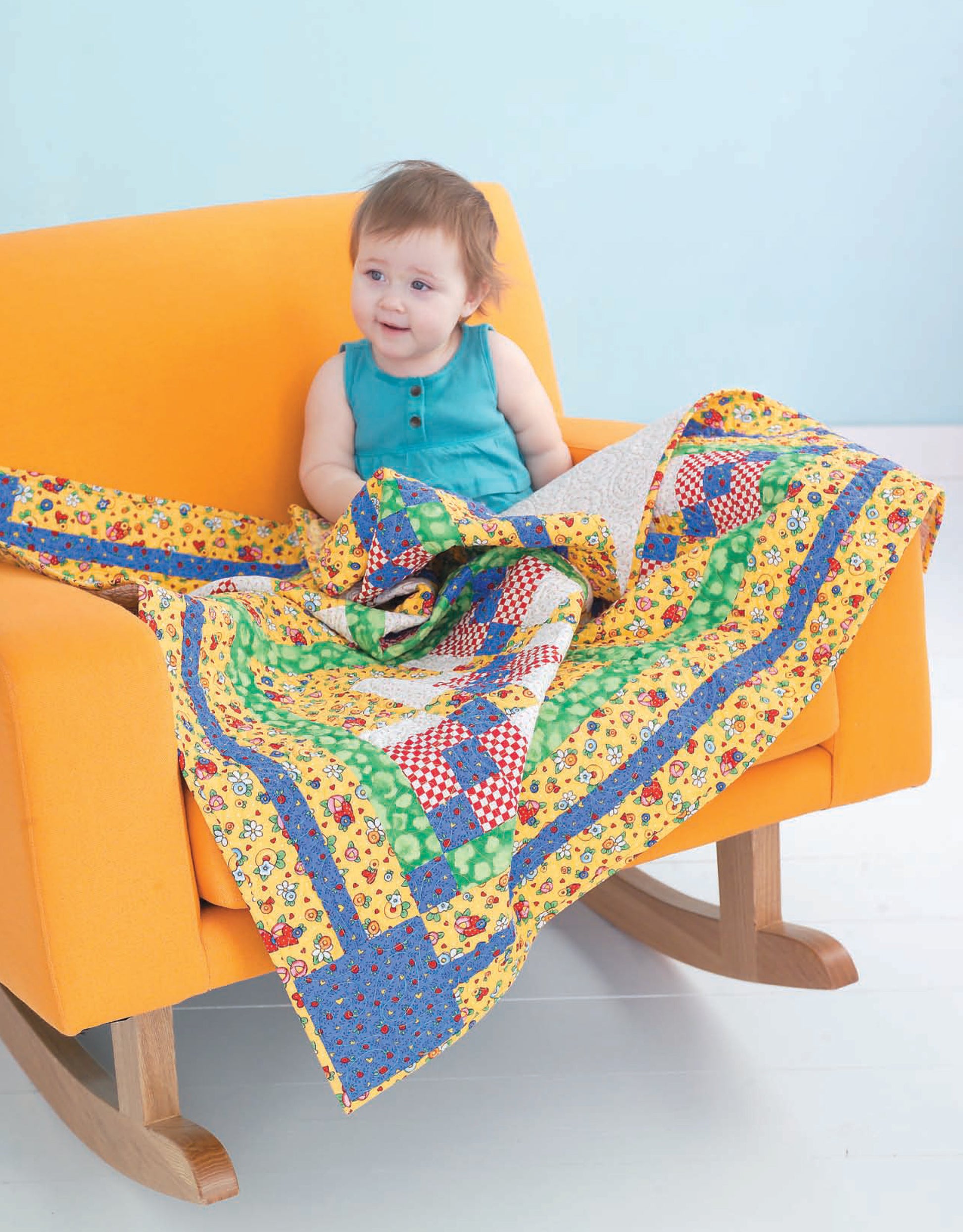 A baby in a turquoise outfit smiles and plays with the colorful Wee Wonder Quilts Digital Download by Leisure Arts while sitting on an orange rocking chair. The backdrop is light blue, and the floor is white.