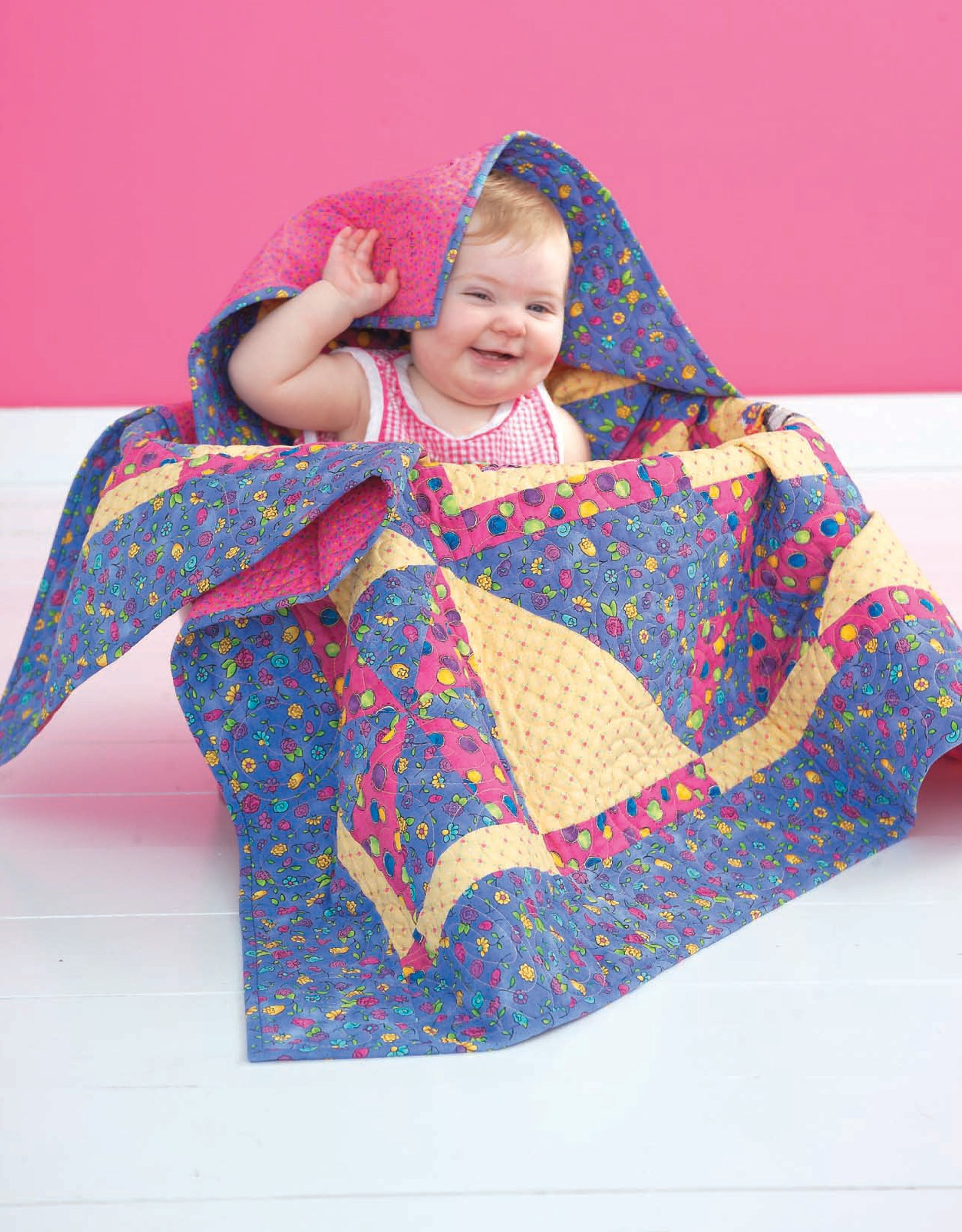 A smiling baby sits on a white floor partially covered by the Leisure Arts "Wee Wonder Quilts" digital download, showcasing playful quilt designs against a bright pink background.