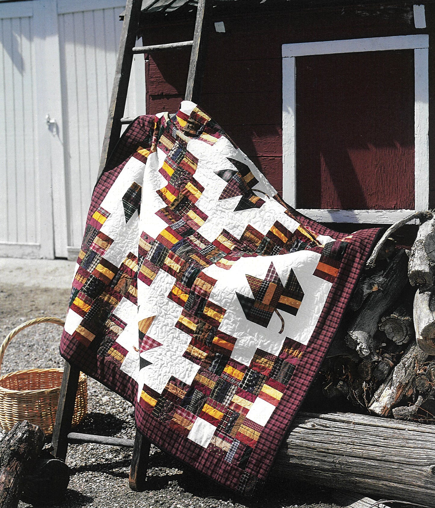 The Quick & Scrappy Quilts by Leisure Arts, featuring vibrant patchwork and leaf designs, is draped over a wooden ladder against a red barn, with logs and a woven basket nearby in the sunny outdoor setting.