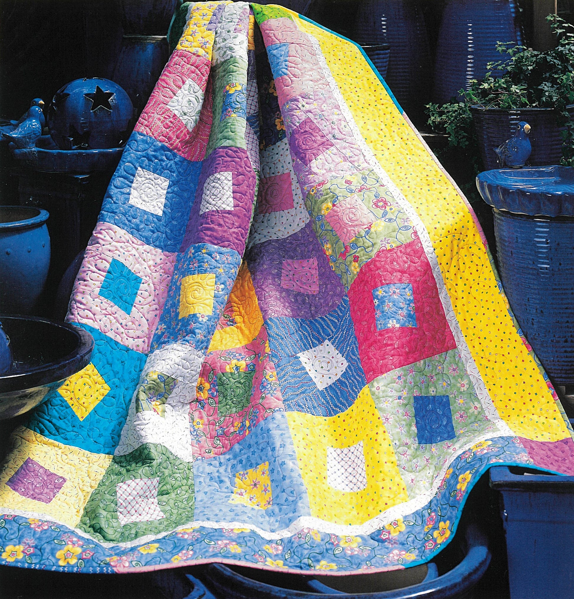 The Quick & Scrappy Quilts by Leisure Arts features a vibrant patchwork of pink, purple, blue, yellow, and green diamonds and squares, displayed with dark blue ceramic pots and plants.