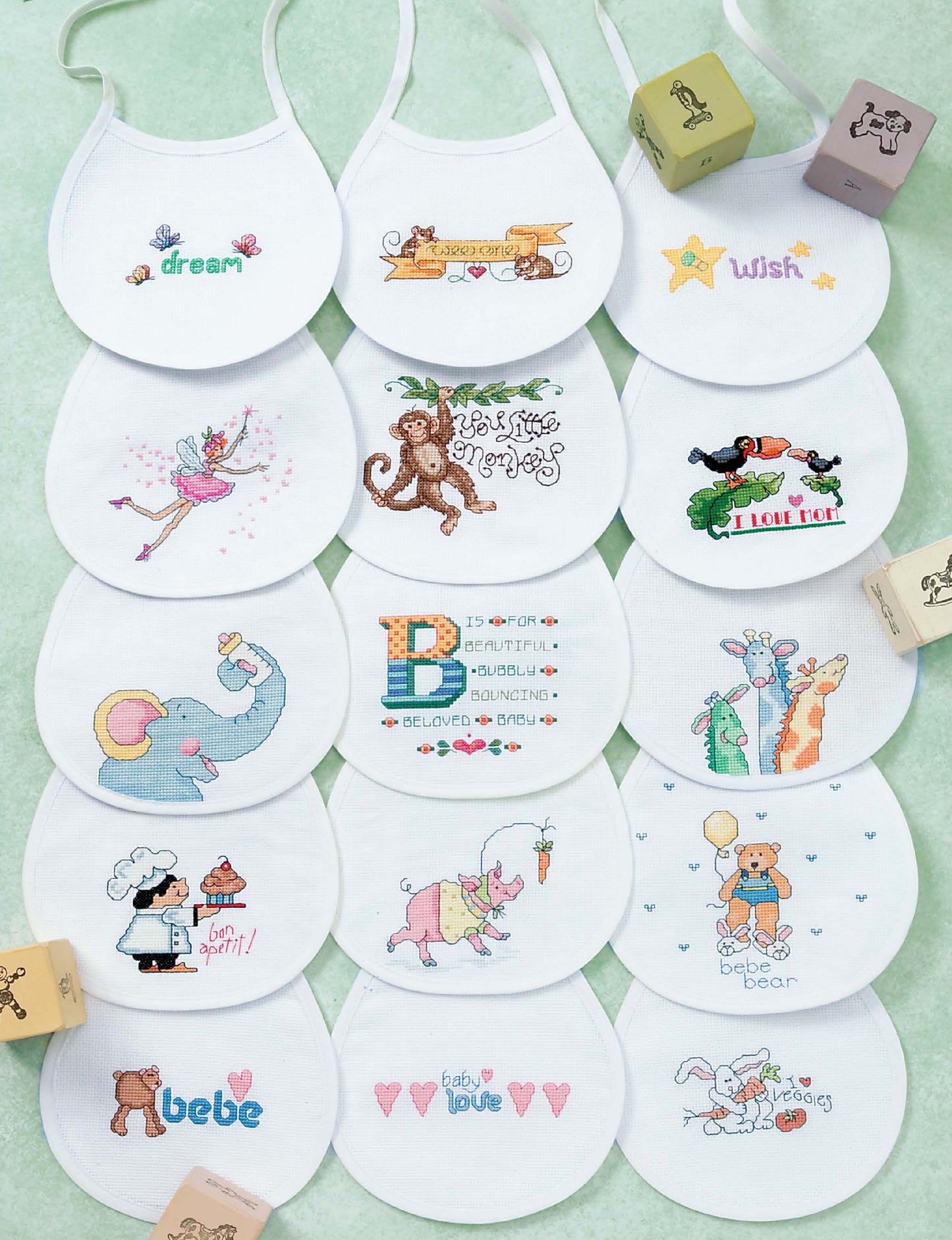 Sixteen white baby bibs from Leisure Arts’ "Baby Bibs to Cross Stitch" are displayed in a grid, each with vibrant embroidered designs—animals, fairies, hearts, and words like “dream” and “wish”—inspired by Linda Gillum’s patterns. Wooden blocks are in the corners.