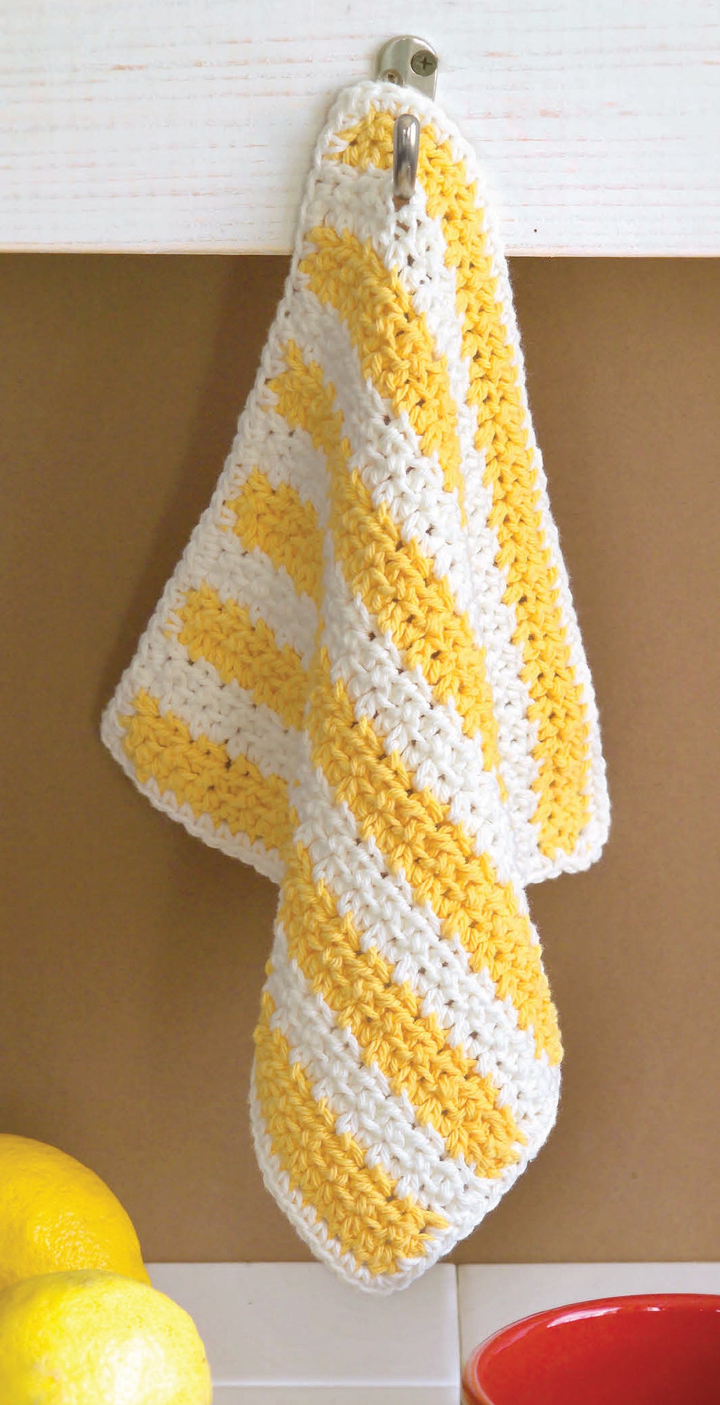 A yellow and white striped crocheted dishcloth hangs from a hook. Perfect for beginners, I Can't Believe I'm Crocheting! by Leisure Arts offers simple instructions. Lemons and a red bowl sit below the board.