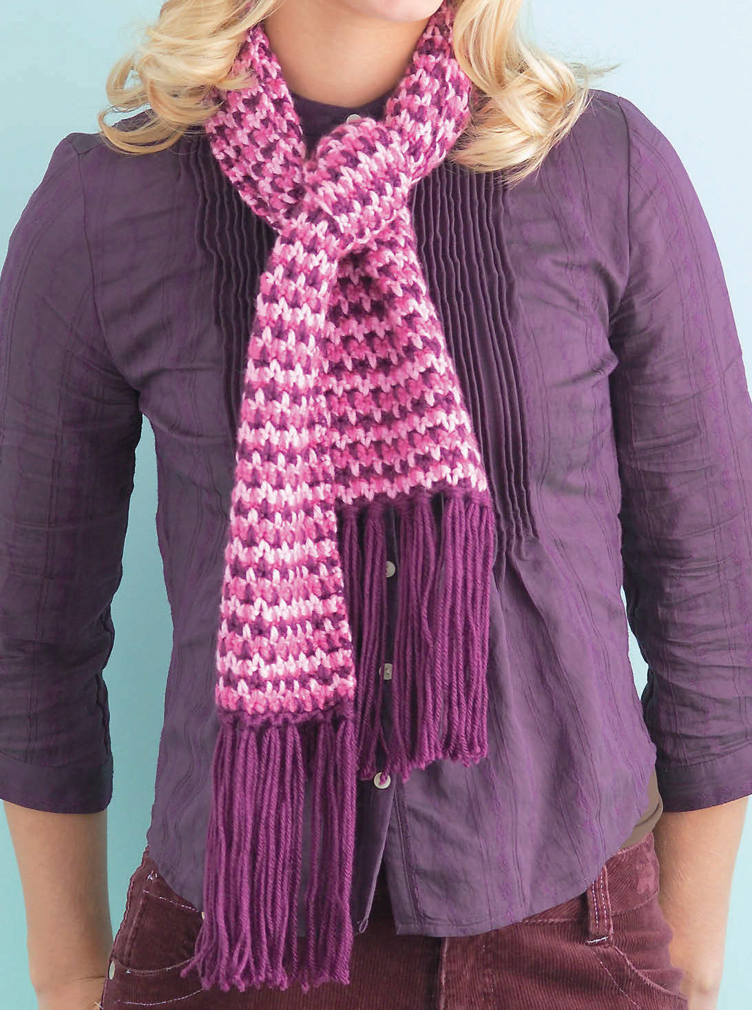 Wearing a purple button-up and a pink and white knit scarf with long purple fringe, this person is ready to follow the “I Can't Believe I'm Crocheting! Digital Download” by Leisure Arts against a light blue background.