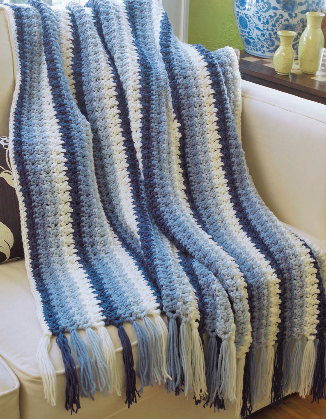 The "I Can't Believe I'm Crocheting!" blanket by Leisure Arts, with thick blue, white, and gray stripes plus long fringe, is draped on a white couch—ideal for beginners and adds cozy charm to any living room.