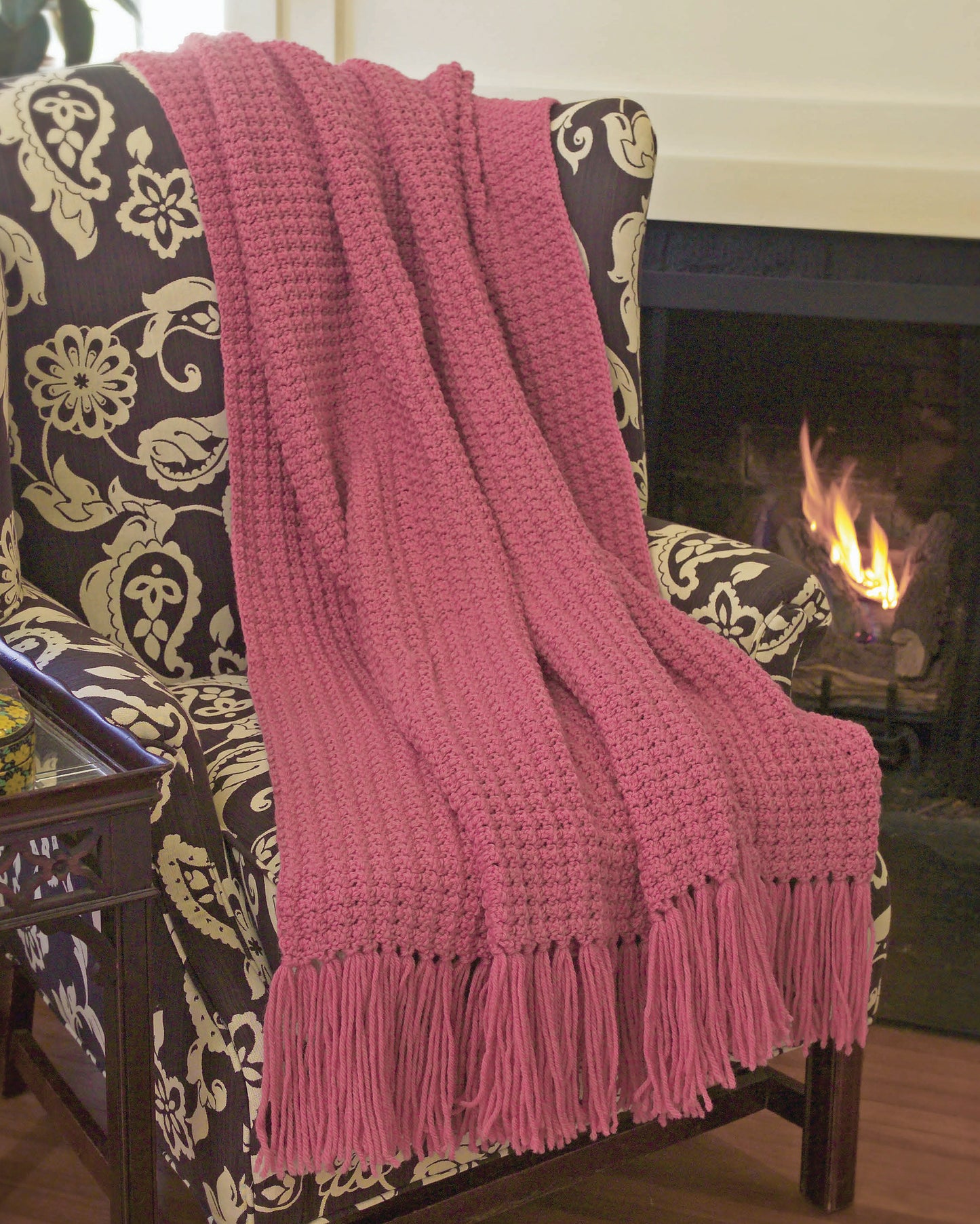A pink knit throw with fringe is draped over a floral armchair by the fire—perfect inspiration for beginners using Leisure Arts' "I Can't Believe I'm Crocheting!" Digital Download.