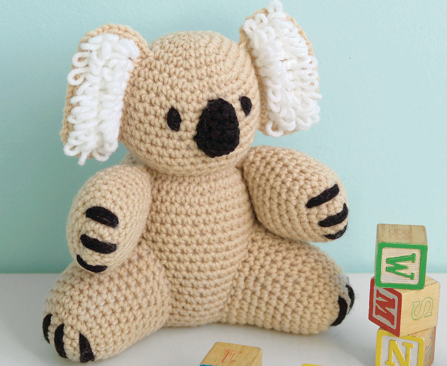 The Leisure Arts "I Can't Believe I'm Crocheting!" book is pictured beside a crocheted koala plush with white ears and black nose, displayed on a white surface near alphabet blocks against a pale blue background.