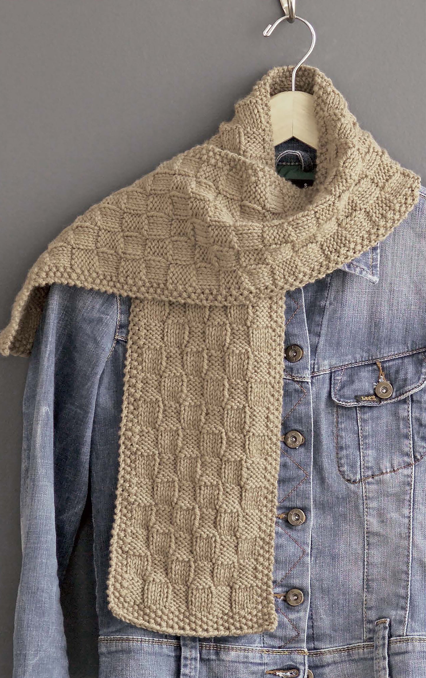 A beige, checkered-knit scarf (ideal for beginners using I Can't Believe I'm Knitting by Leisure Arts) is draped over a blue denim jacket hanging on a hook against a gray wall.