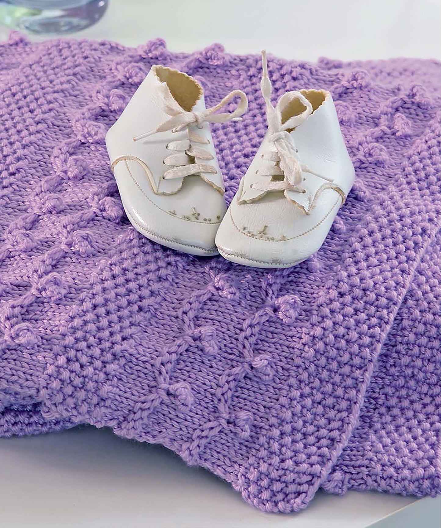 A pair of white baby shoes with laces sits on a soft lavender knitted baby blanket—an adorable project made from the beginner-friendly "I Can't Believe I'm Knitting" guide by Leisure Arts.