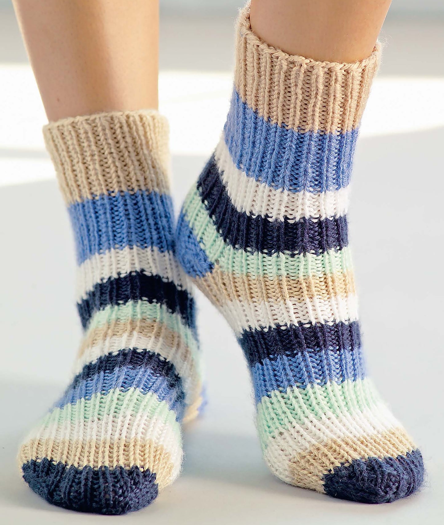 Two feet in thickly striped socks—blue, navy, light green, tan, and cream—stand on a light background. Get inspired for your next project with "I Can't Believe I'm Knitting" from Leisure Arts.