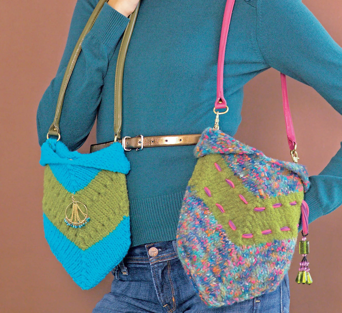 A person in a teal sweater and jeans models two colorful knit shoulder bags made with I Can't Believe I'm Knitting by Leisure Arts: one blue-green with a gold charm, the other green with multicolored yarn and pink stitching. Perfect knitting accessories!.