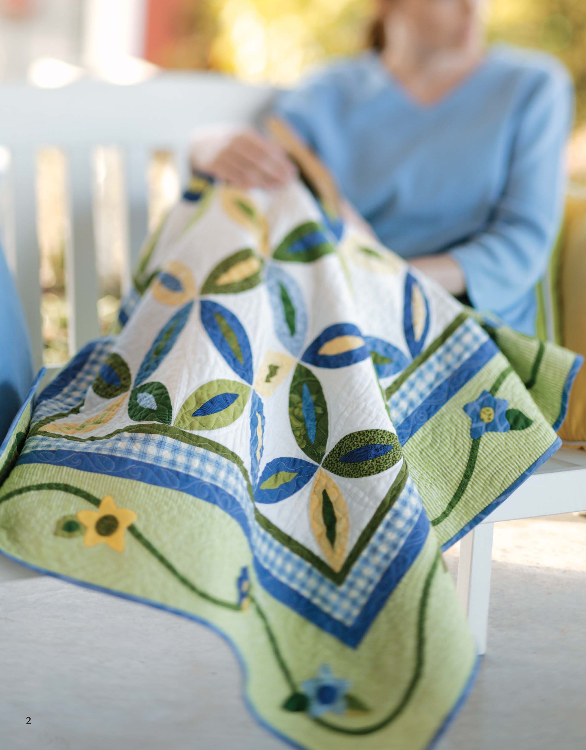 A person in a blue top sits on a bench, holding the "Garden of Appliqué Digital Download" quilt by Leisure Arts, featuring green, yellow, and blue floral and geometric applique designs and a checkered border.