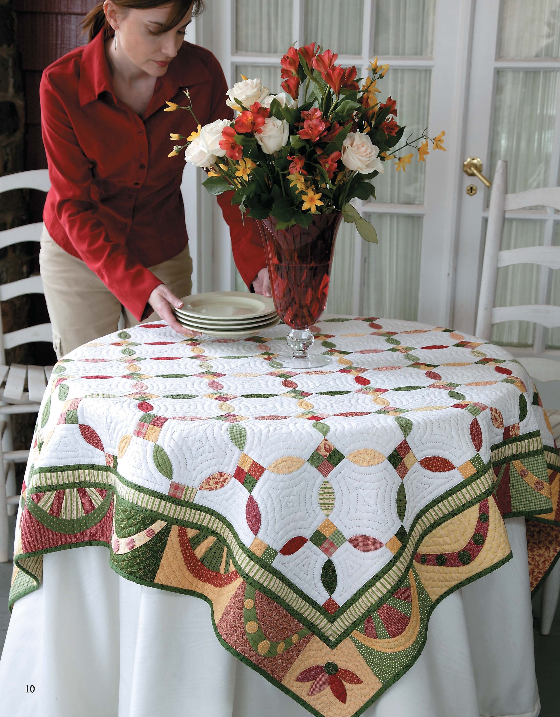 A woman in a red shirt sets plates on a round table covered with the colorful, nature-inspired "Garden of Appliqué Digital Download" quilt by Leisure Arts. A vase of orange, white, and yellow flowers sits at the center, with French doors in the background.