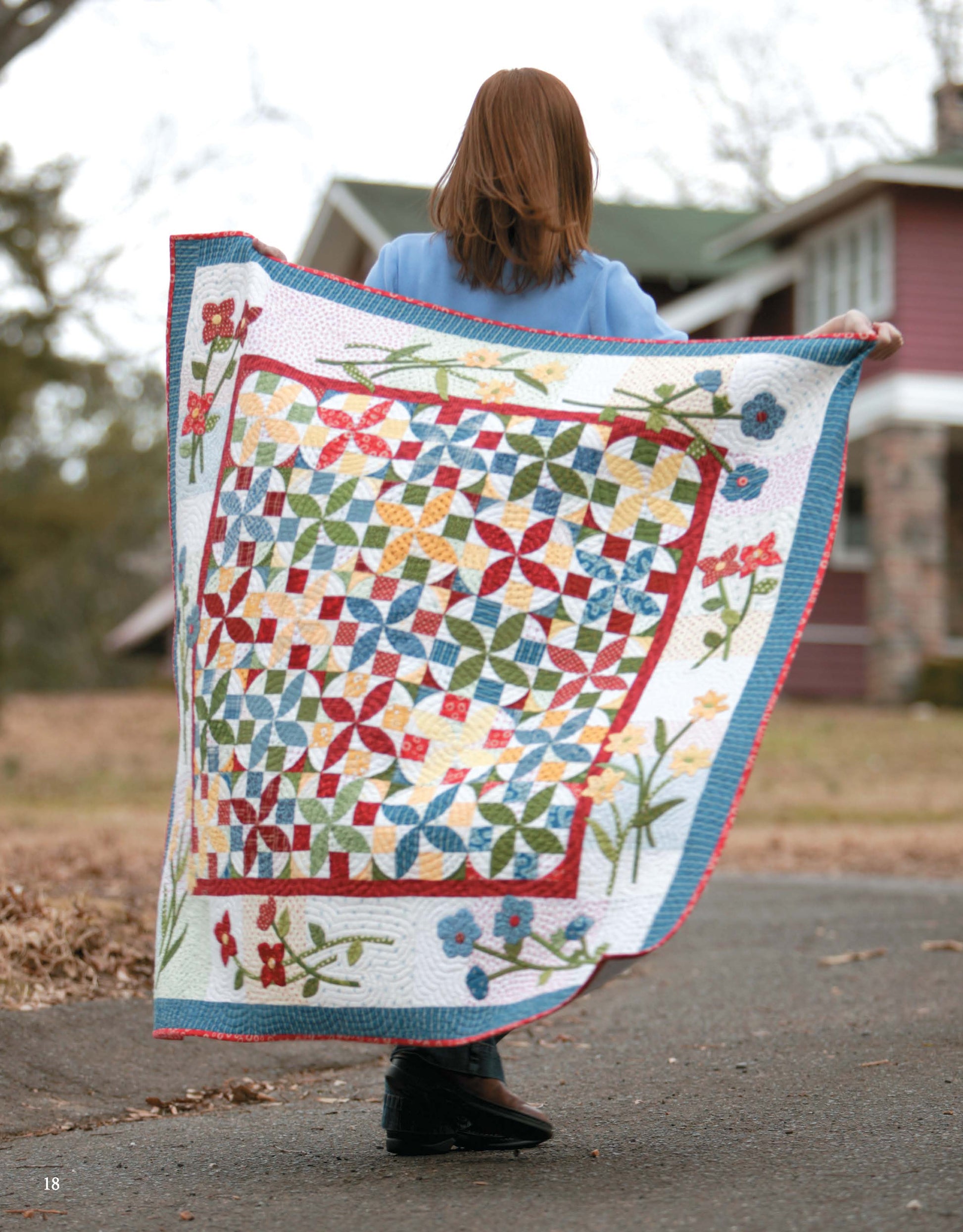 A person stands outside with their back to the camera, holding Leisure Arts’ Garden of Appliqué Digital Download quilt featuring floral patterns; a house and trees in the background capture the essence of nature-inspired quilting.