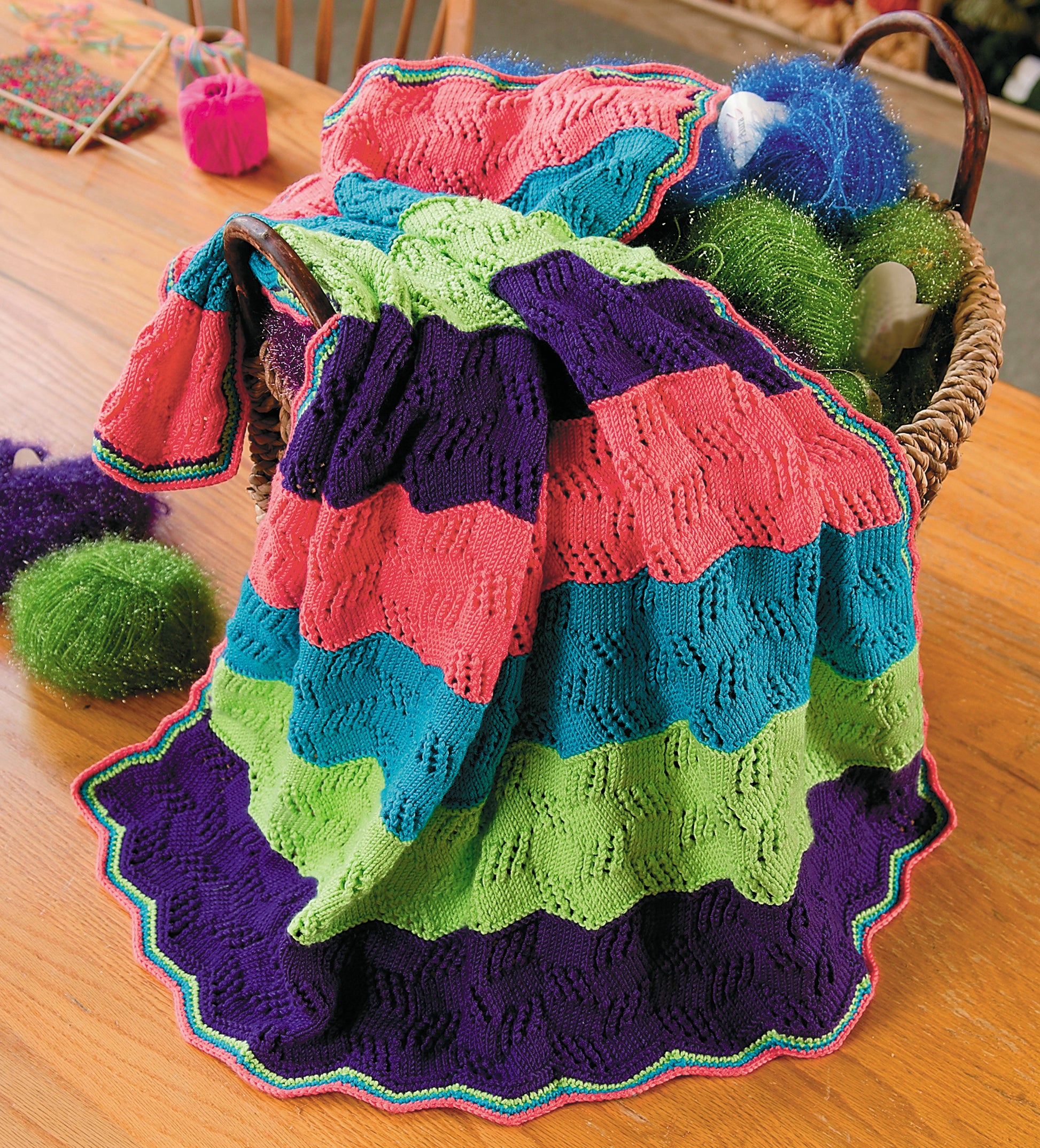 A vibrant, wavy-patterned baby afghan in pink, green, blue, and purple drapes from a wicker basket on a wooden table with yarn balls—a cozy project from Knit Along with Debbie Macomber—The Shop on Blossom Street by Leisure Arts.