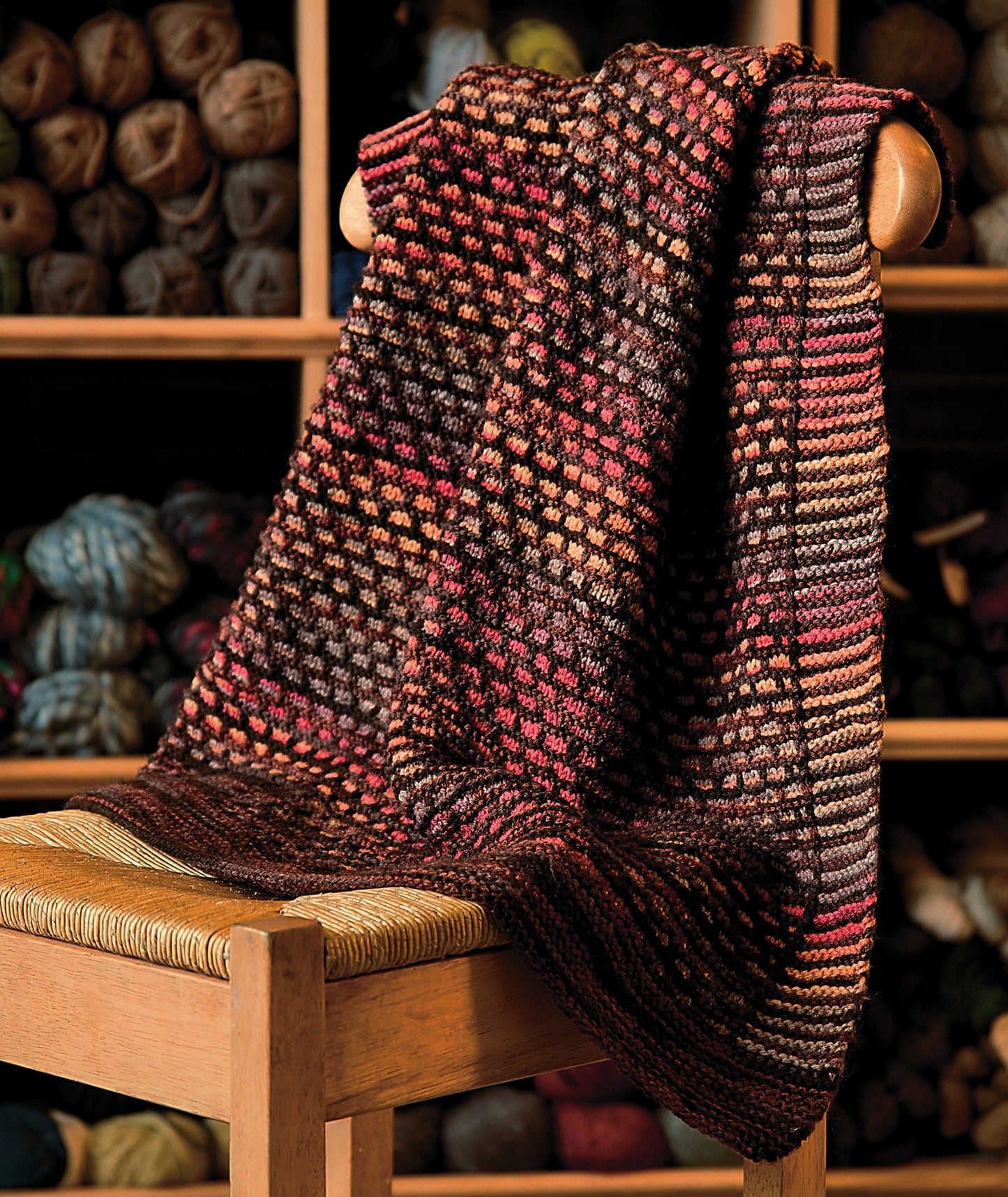 A multicolored knitted blanket in red, brown, and orange drapes over a wooden chair. Behind it, shelves hold colorful yarn balls—an inspiring scene for fans of *Knit Along with Debbie Macomber—The Shop on Blossom Street* by Leisure Arts.