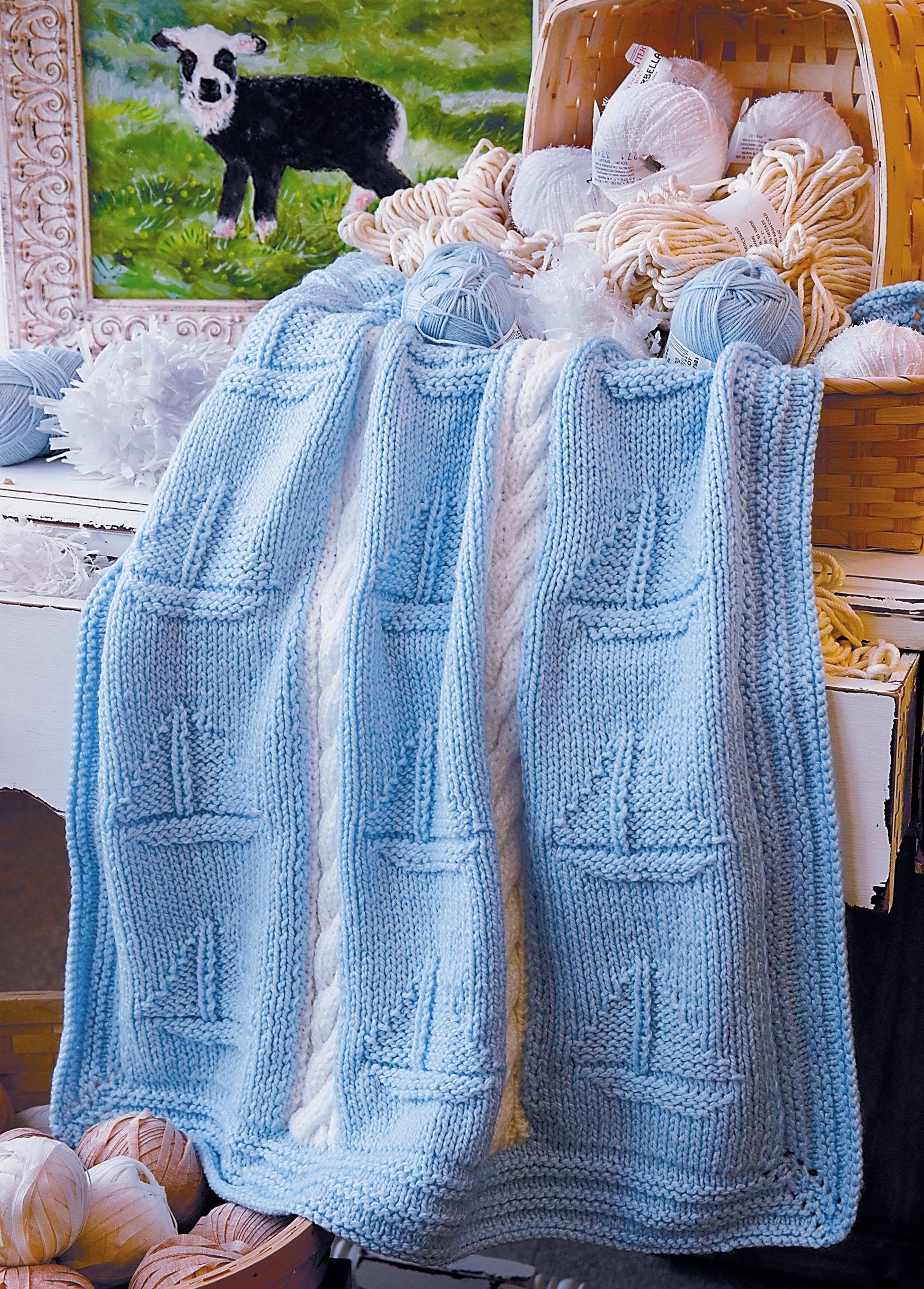 A light blue baby blanket from Leisure Arts’ “Knit Along with Debbie Macomber—The Shop on Blossom Street,” featuring white cables and sailboats, drapes over a wicker basket of pastel yarn. A lamb painting is in the background.