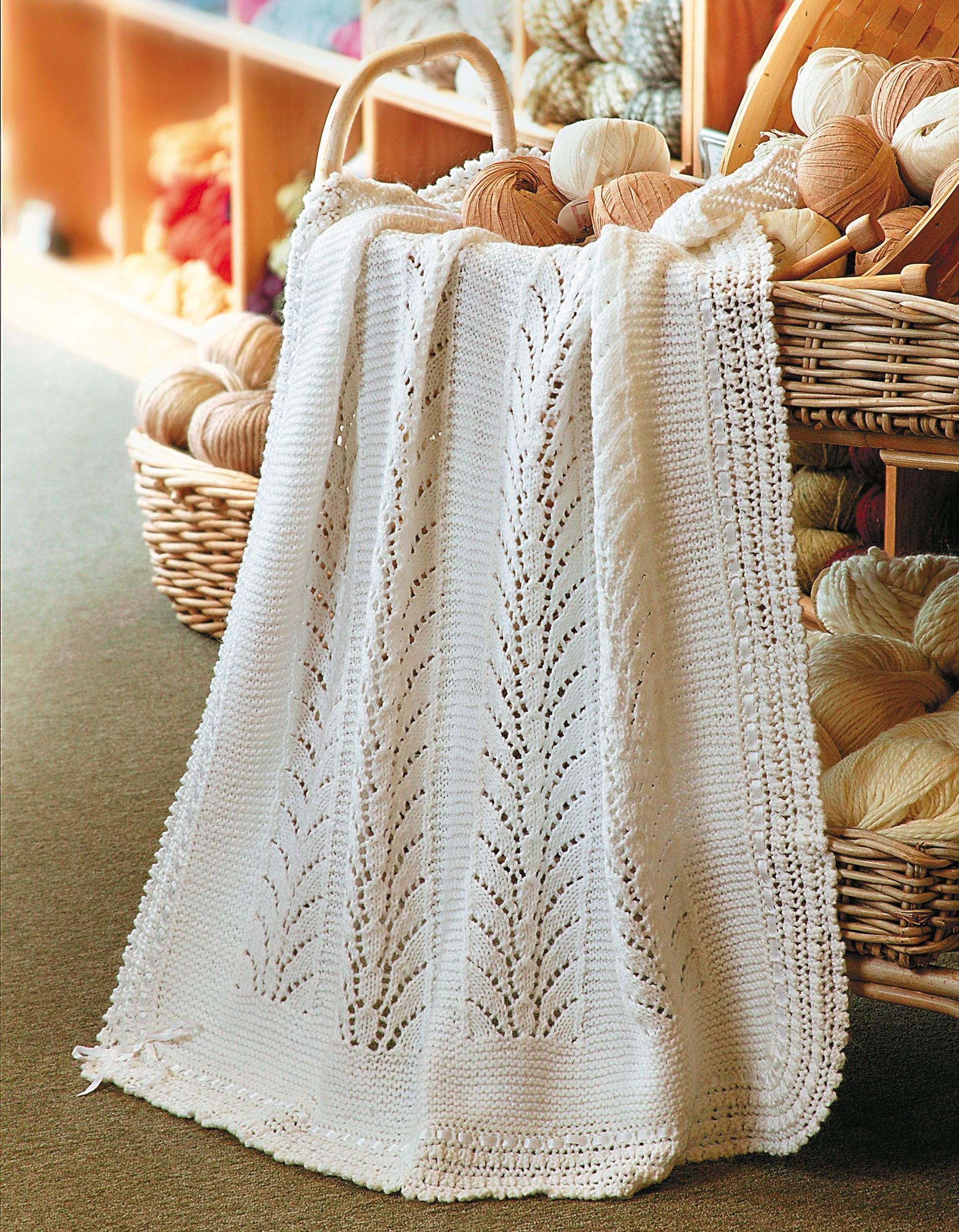 A white, intricately knit leaf-pattern blanket from “Knit Along with Debbie Macomber—The Shop on Blossom Street” by Leisure Arts is draped over a woven basket of yarn in a cozy craft room.