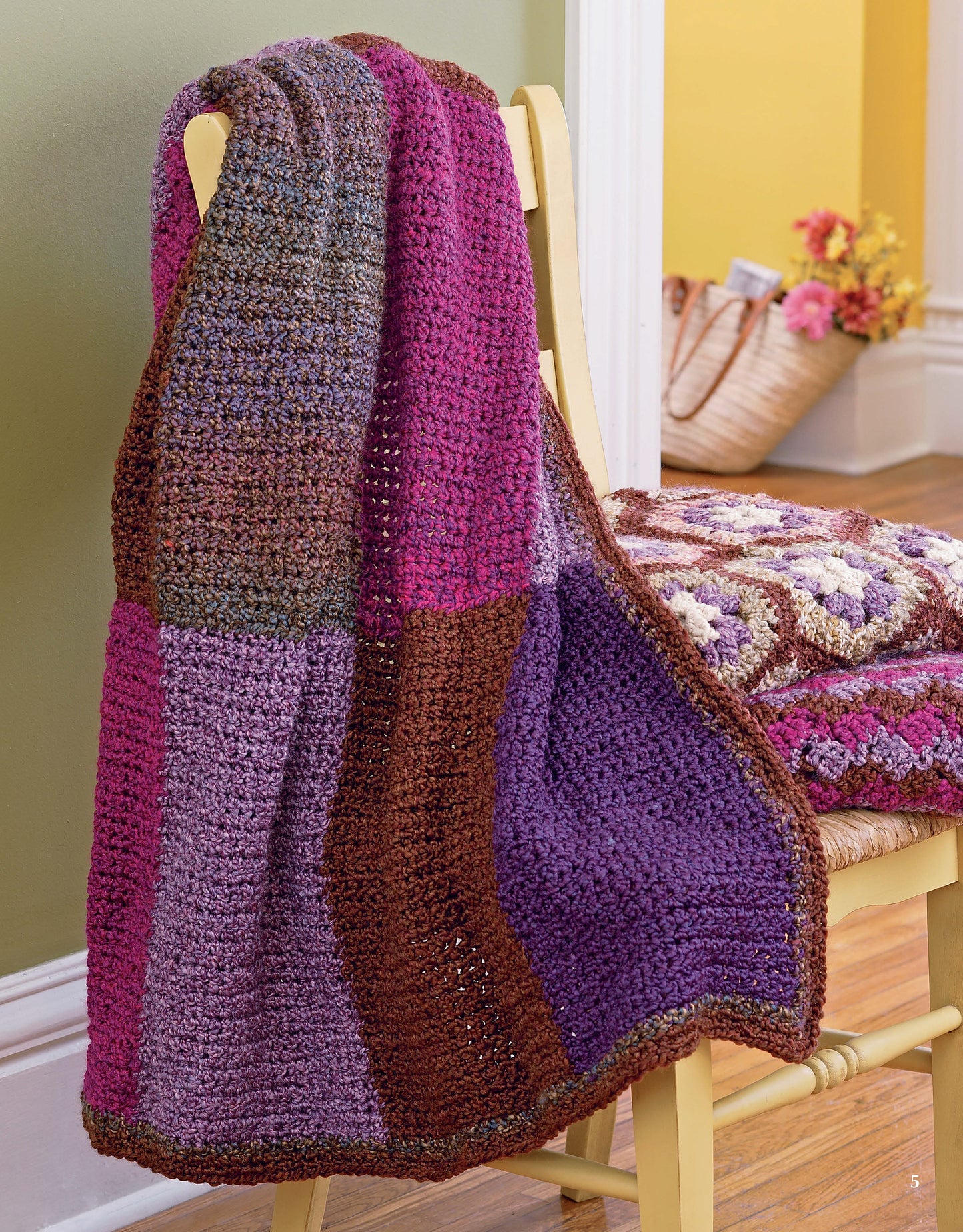 A cozy blanket from Leisure Arts’ Homespun Afghans, featuring purple, pink, and brown hues, is draped over a yellow chair. A basket of flowers and another easy crochet throw rest on the wooden floor by the green and yellow wall.