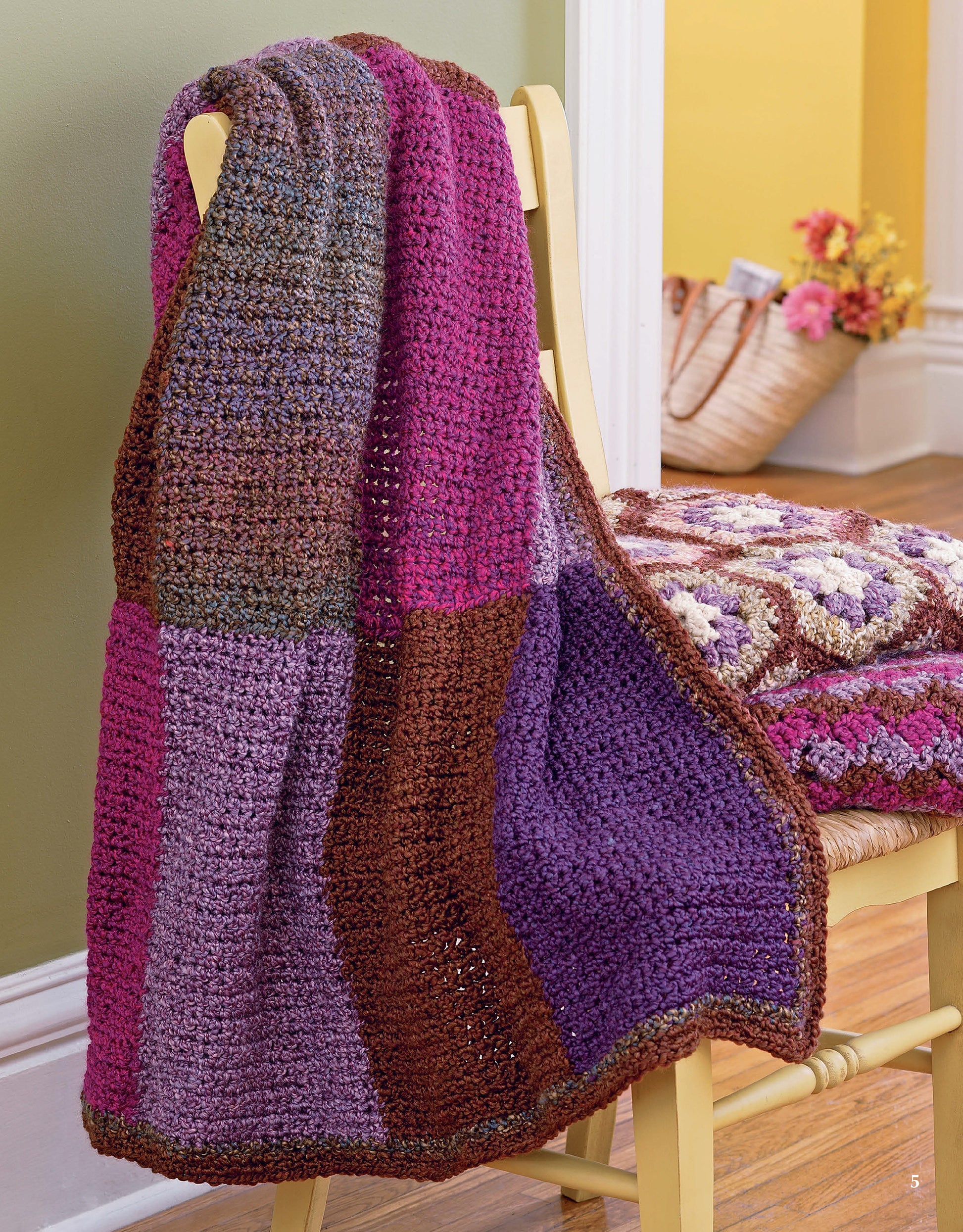 A cozy blanket from Leisure Arts’ Homespun Afghans, featuring purple, pink, and brown hues, is draped over a yellow chair. A basket of flowers and another easy crochet throw rest on the wooden floor by the green and yellow wall.