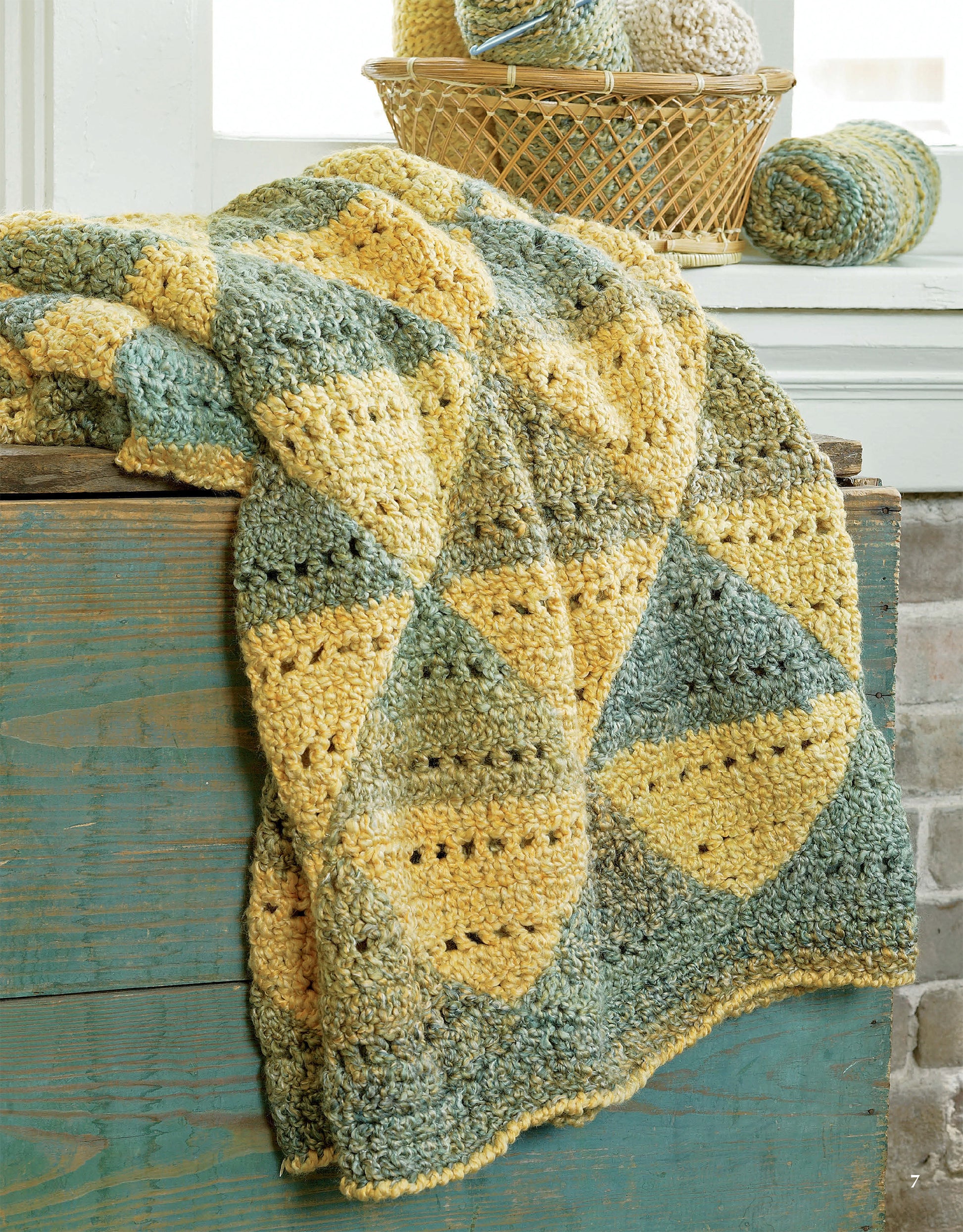 A yellow and green crocheted blanket from Homespun Afghans by Leisure Arts drapes over a rustic blue chest, while a basket of matching yarn sits by the window.
