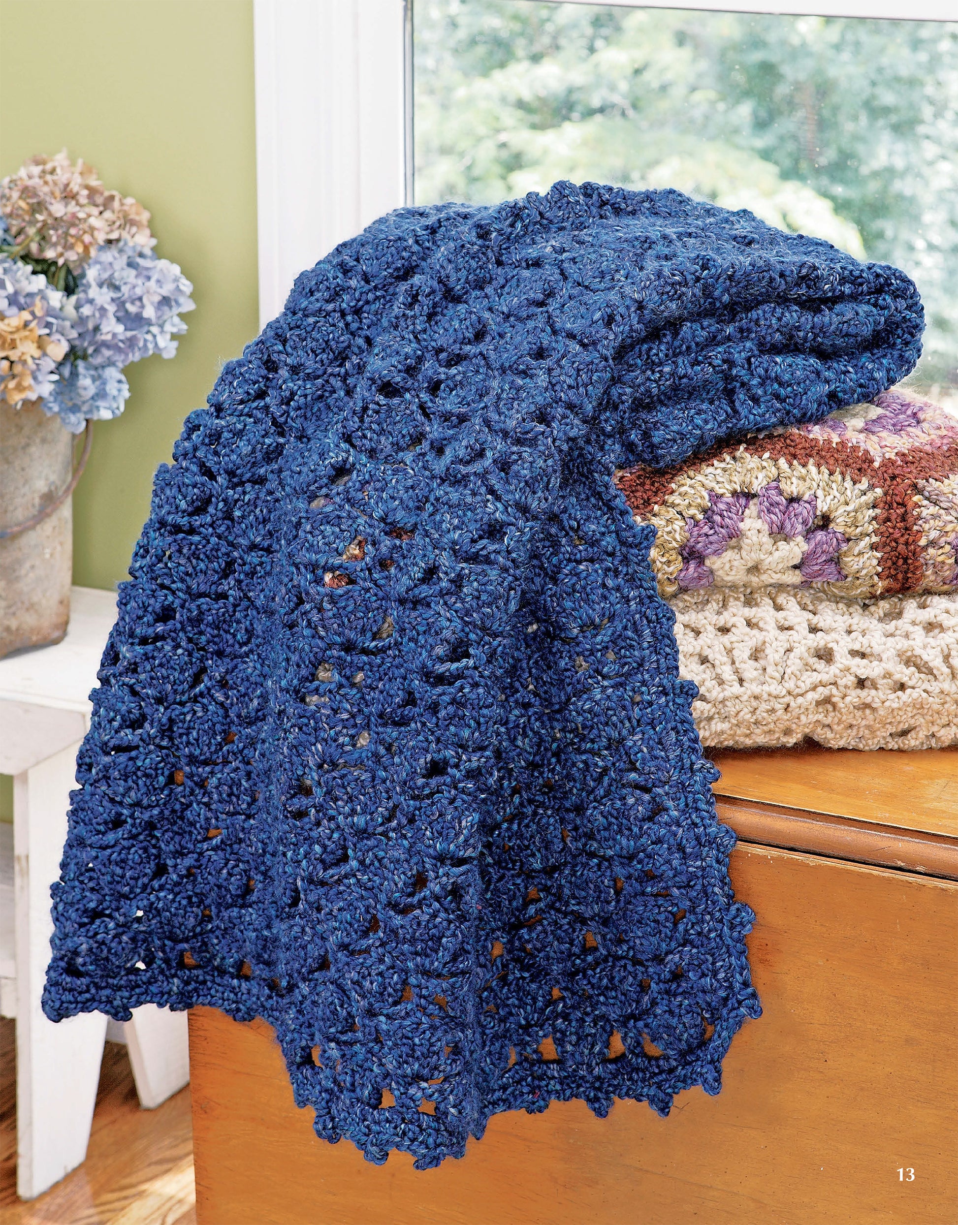 A textured dark blue blanket from Homespun Afghans by Leisure Arts is draped over a wooden chair near a window, with a vase of dried hydrangeas and folded blankets in the background.