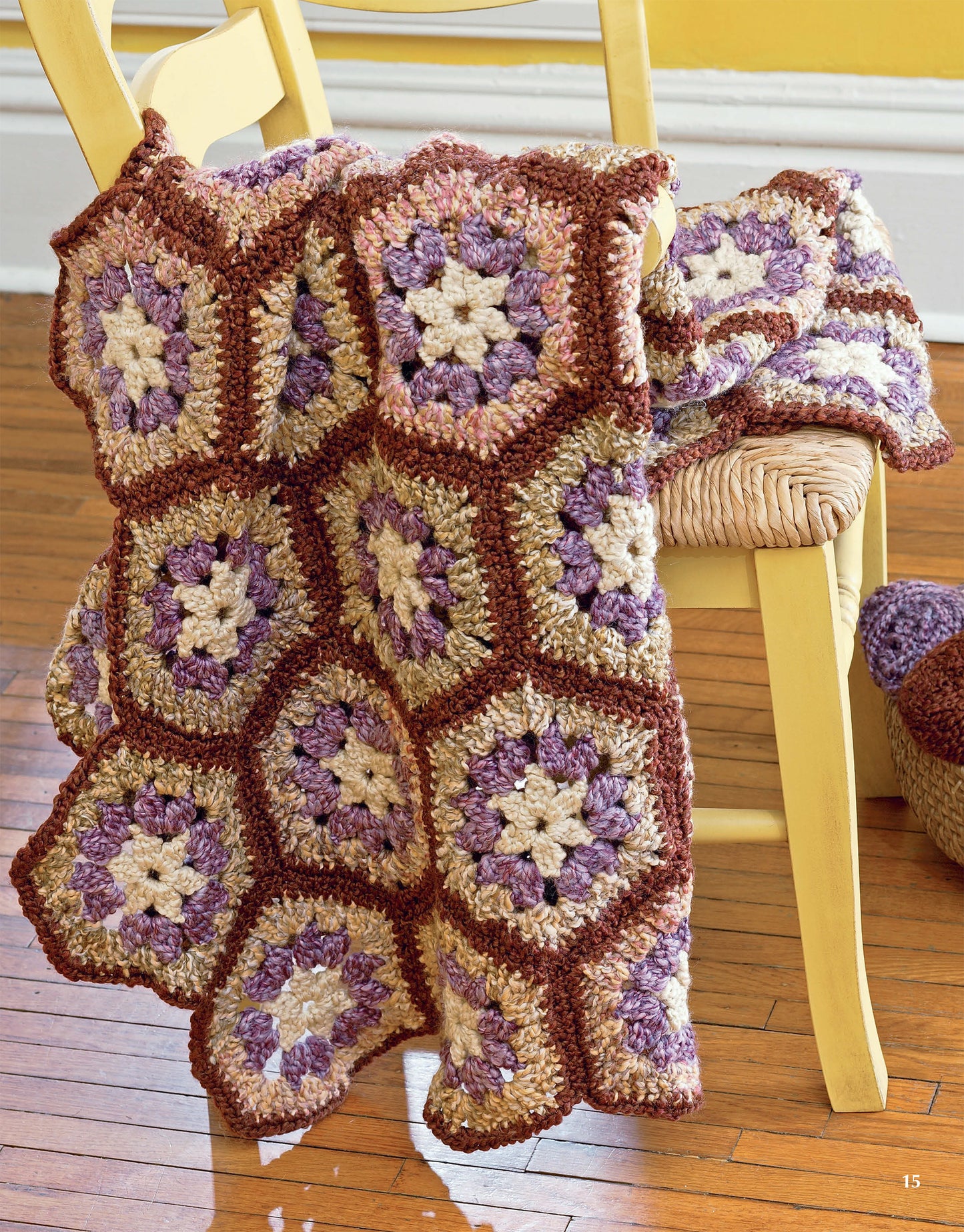 The Homespun Afghans from Leisure Arts, featuring hexagonal crochet motifs in brown, cream, and purple hues, is draped over a light yellow wooden chair on a polished hardwood floor.
