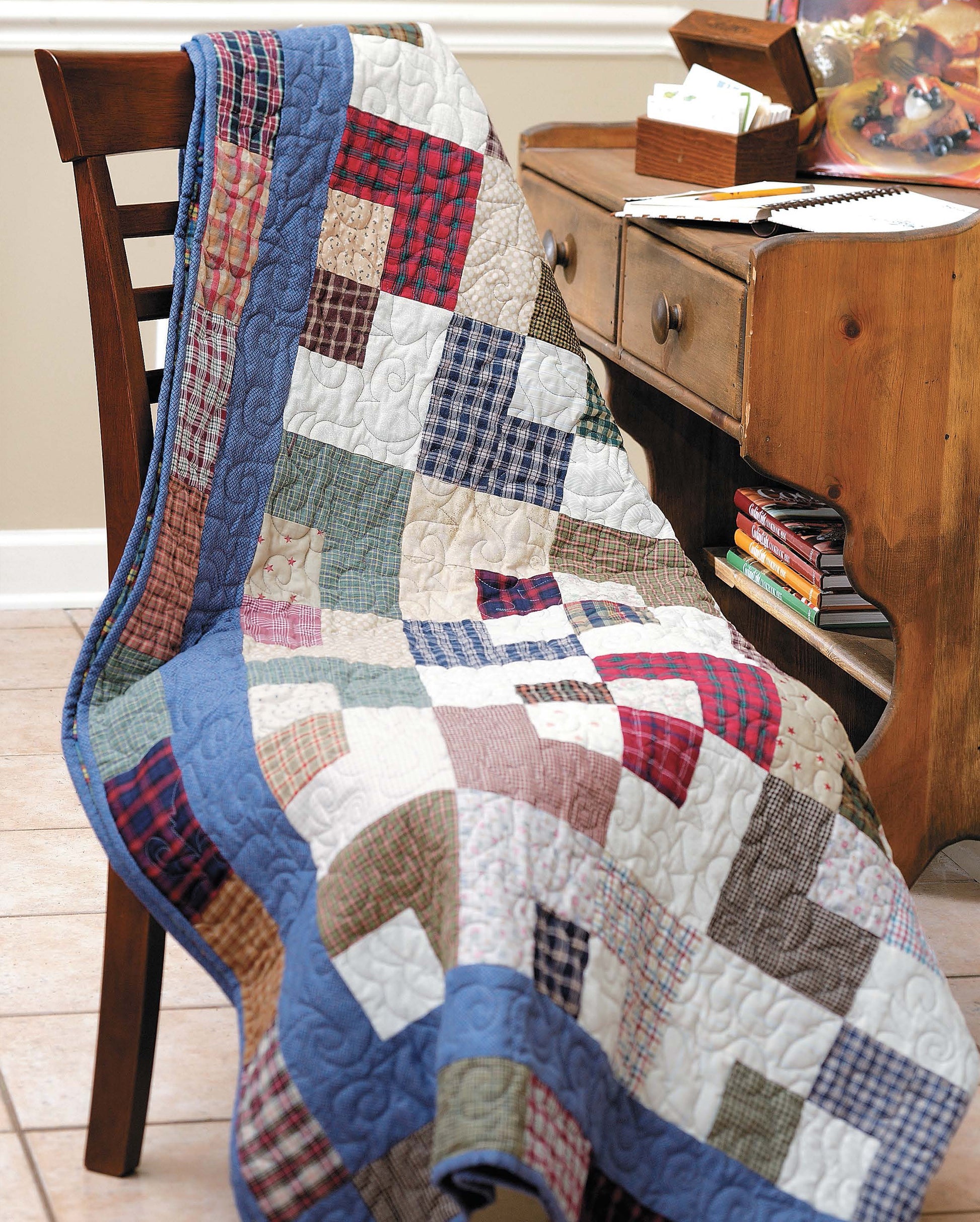 The Slash Your Stash Quilts by Leisure Arts, crafted with fast piecing, features a blue border. It's shown draped over a wooden chair next to a desk with books and art supplies in a room with cream walls and tiled floors.