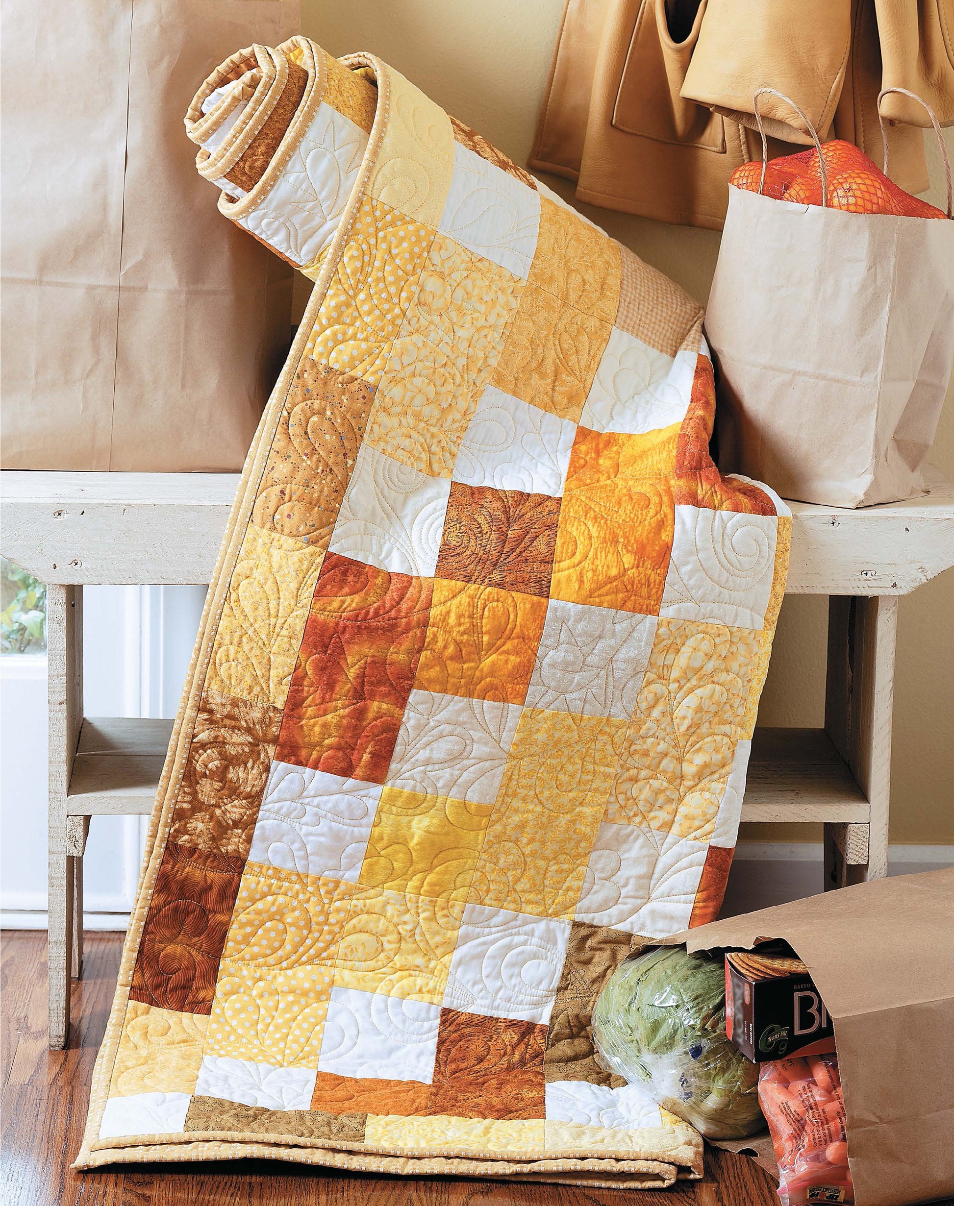 The Slash Your Stash Quilts by Leisure Arts, in yellow, orange, and white patchwork made with fast piecing techniques, is draped over a bench beside brown grocery bags filled with lettuce and oranges on a wooden floor.