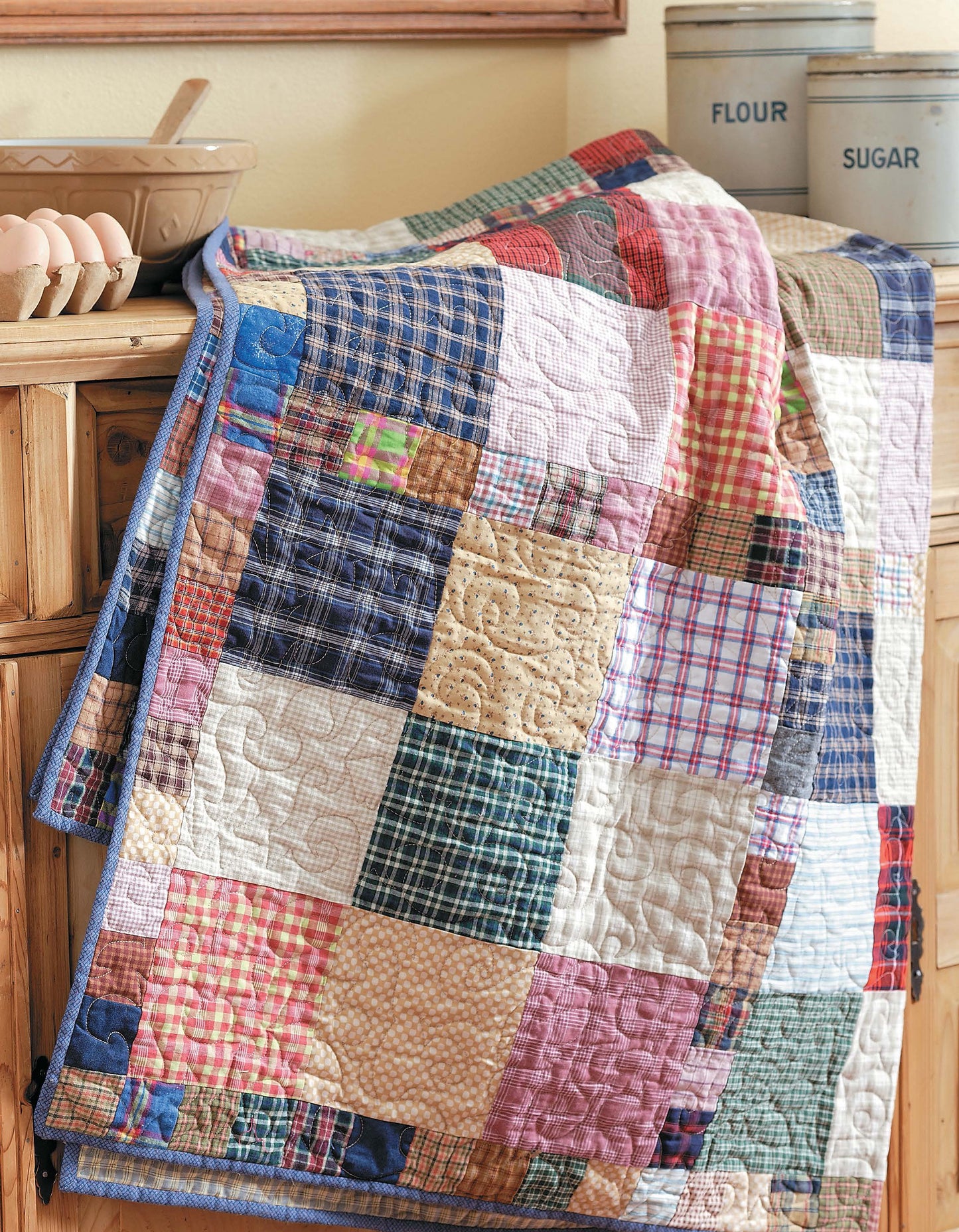 The Slash Your Stash Quilts by Leisure Arts features a vibrant patchwork design made with speedy piecing to minimize your stash. Displayed over a cabinet, it’s paired with vintage flour and sugar canisters and a bowl of eggs for nostalgic appeal.