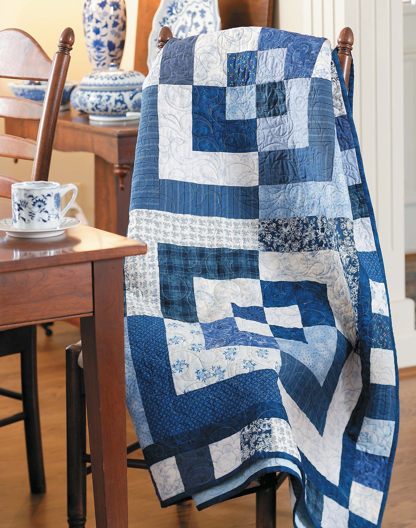 The Slash Your Stash Quilts by Leisure Arts, in blue and white patchwork, is draped over a wooden chair beside a table with a matching teacup and blue and white ceramic vases.