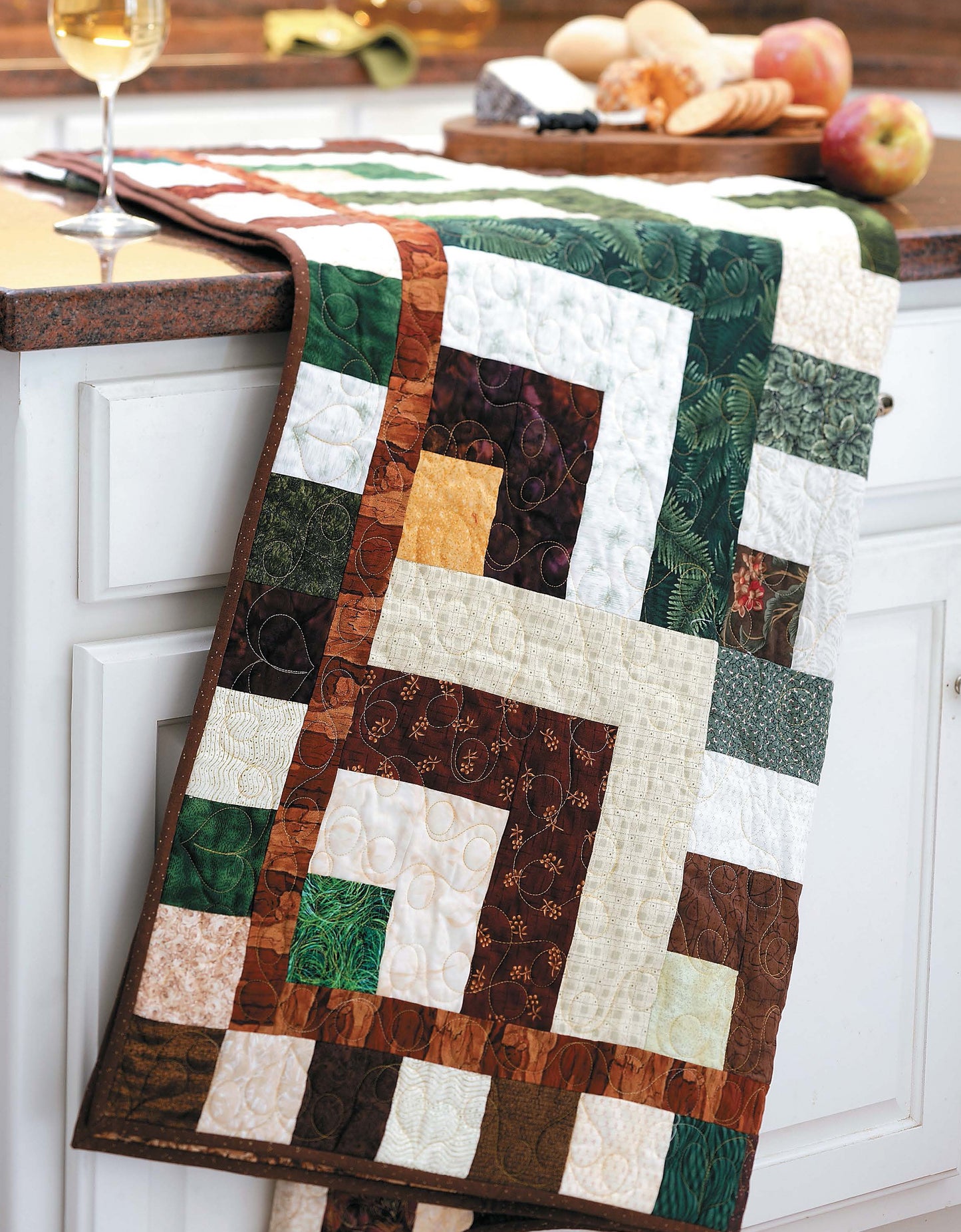 The Slash Your Stash Quilts runner by Leisure Arts, crafted with quick piecing to use up your fabric stash, is draped on a white kitchen counter with apples, bread, cheese, and a glass of white wine in the background.