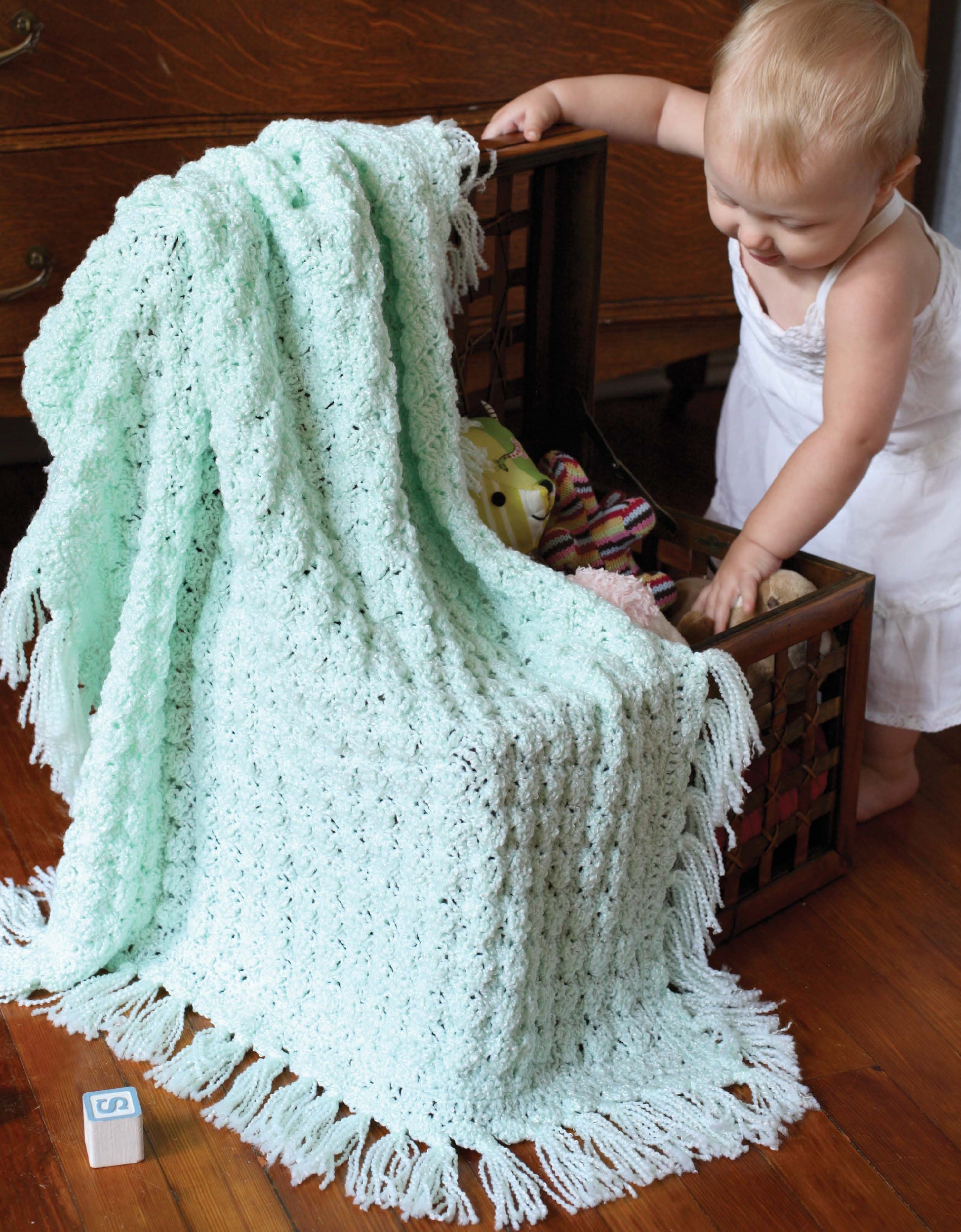 A baby in a white dress stands by an open toy chest draped with a textured, pale blue afghan made from Crochet Classics for Baby by Leisure Arts, as a toy block with the letter S rests on the hardwood floor nearby.