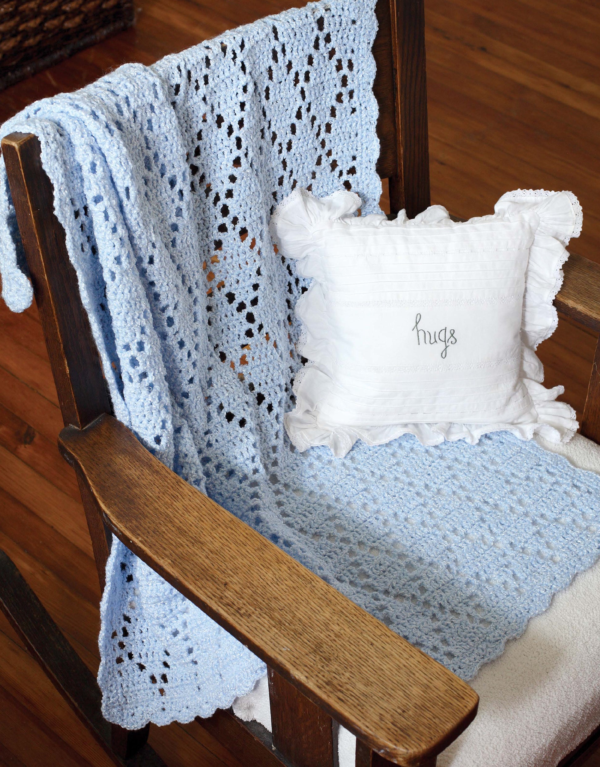 A wooden rocking chair draped with a light blue blanket made using Leisure Arts’ "Crochet Classics for Baby," perfect for beginner projects, and a white ruffled pillow with "hugs" on it sits on a wooden floor.