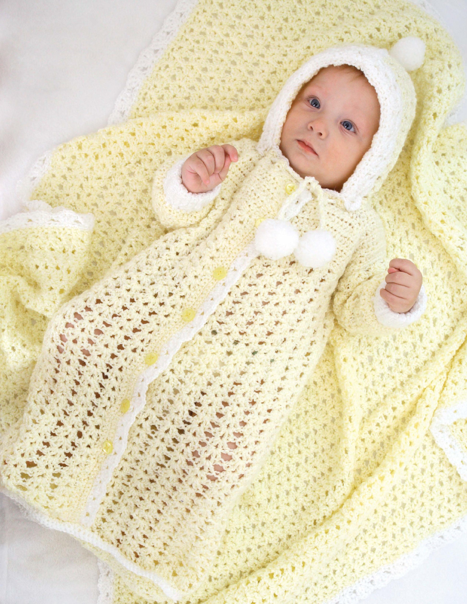 A baby lies on a yellow crocheted blanket, wearing a matching yellow and white hooded gown with two pom-poms—just one of the adorable beginner-friendly patterns from Crochet Classics for Baby by Leisure Arts. The baby gazes up calmly.