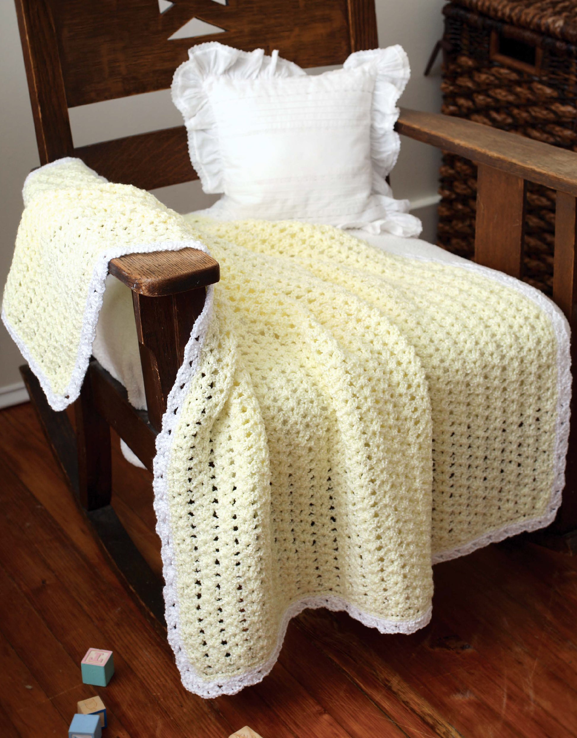 A pale yellow blanket from "Crochet Classics for Baby" by Leisure Arts, featuring a white scalloped border, is draped over a wooden rocking chair with a white pillow on a hardwood floor beside wicker baskets and colorful toy blocks.
