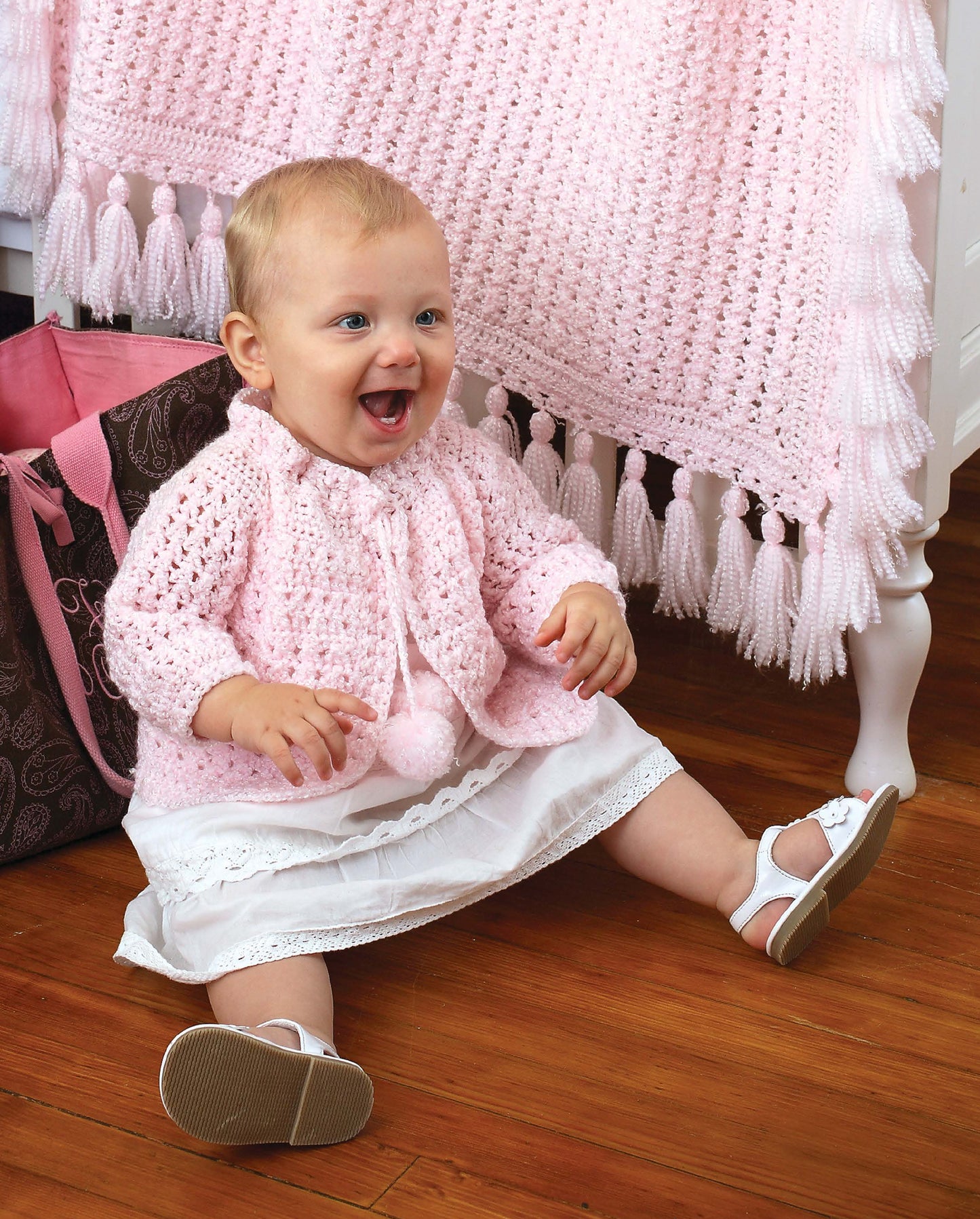A smiling baby in a pink crocheted sweater and white dress sits on a wooden floor with a brown and pink bag beside her, showcasing sweet designs from Leisure Arts’ Crochet Classics for Baby—perfect beginner patterns for adorable baby projects.