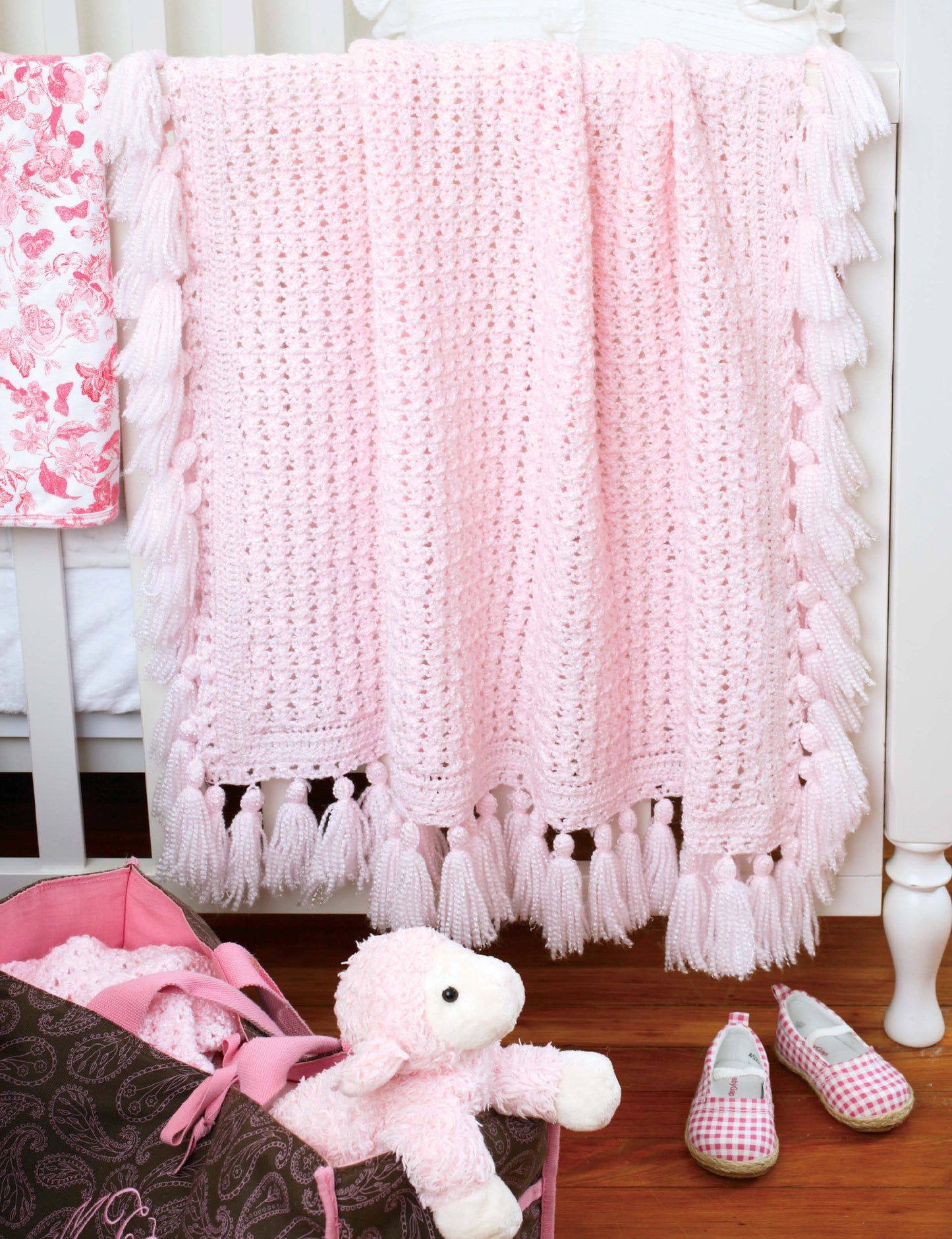 A soft pink baby blanket made with Leisure Arts' Crochet Classics for Baby hangs on a white crib. On the floor beneath are a plush lamb toy, pink and brown diaper bag, and pink plaid baby shoes.