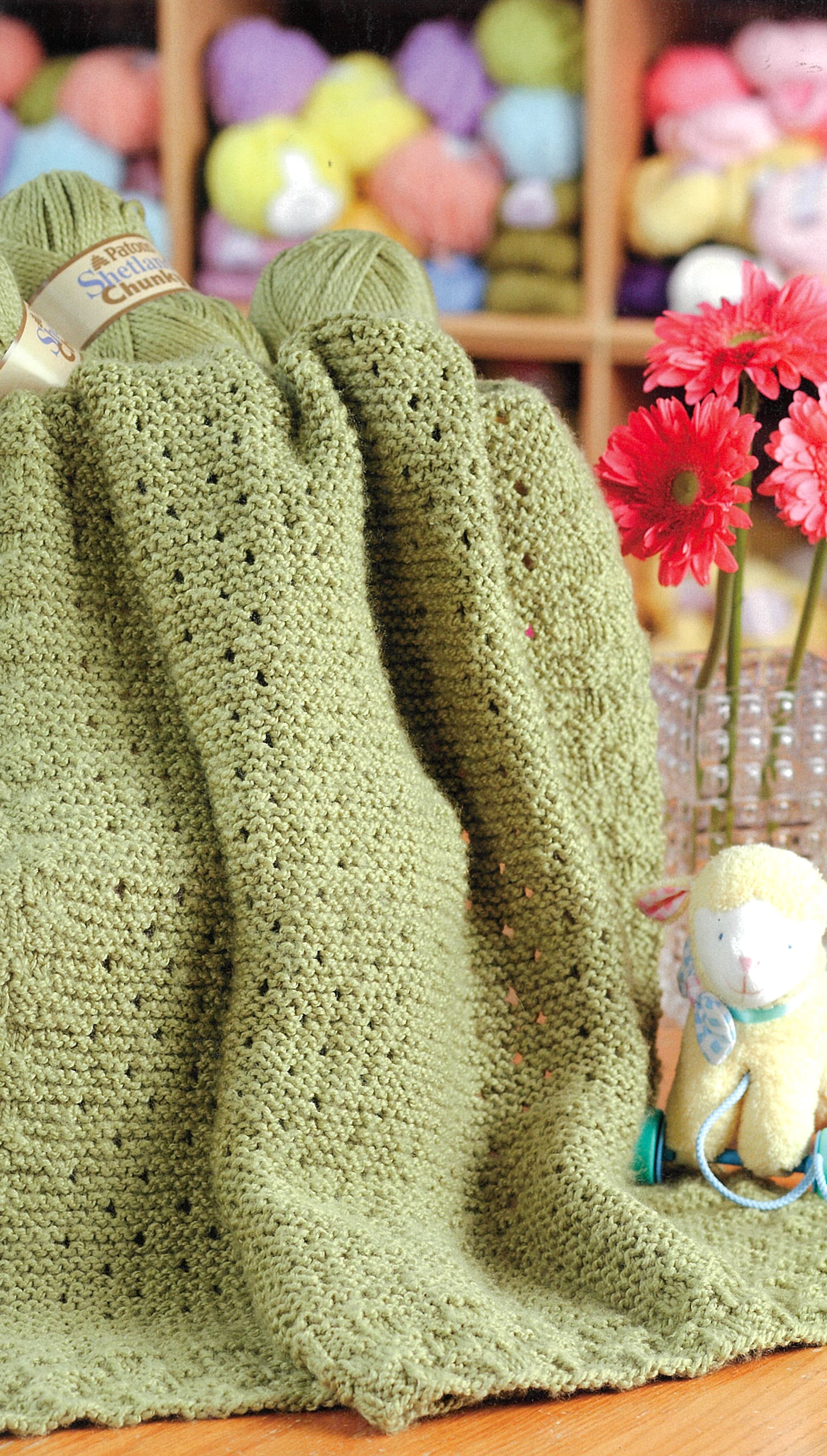 A green knitted blanket drapes over a table with yarn balls and red flowers nearby. A stuffed animal sits by shelves of colorful yarn—inspiration for Leisure Arts’ Knit Along with Debbie Macomber—Back on Blossom Street.
