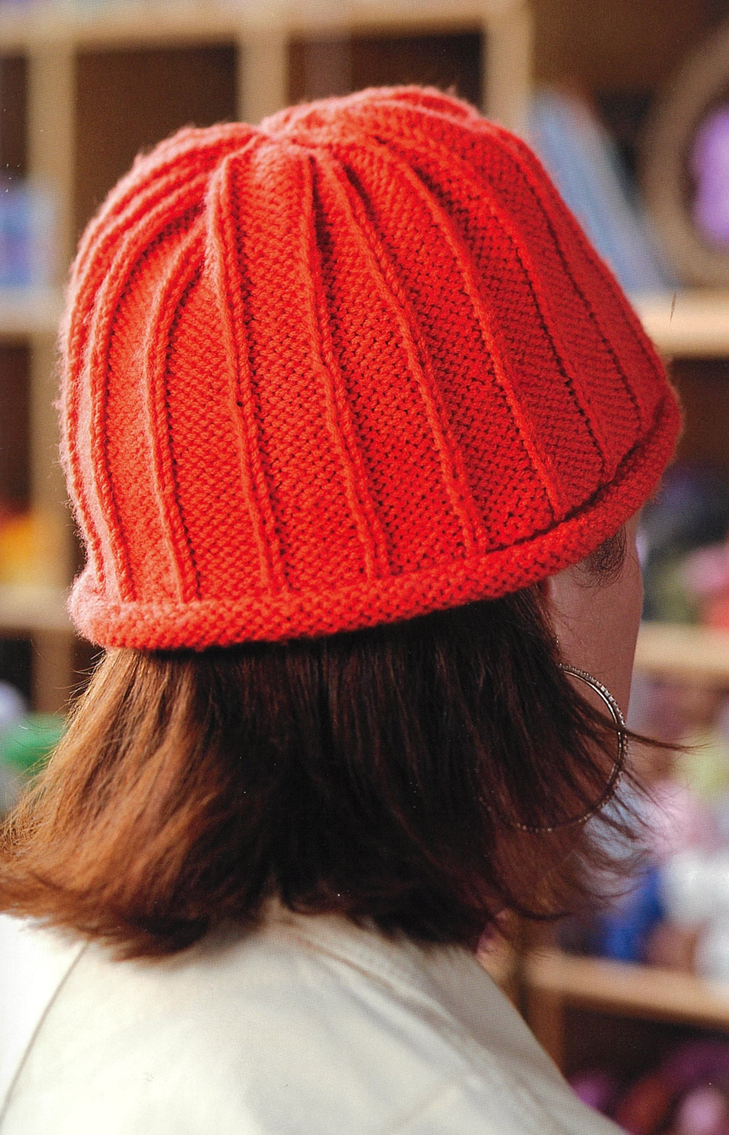 A person with shoulder-length brown hair wears a bright red, vertically ribbed knitted beanie. The photo, taken from behind, shows softly blurred shelves featuring Knit Along with Debbie Macomber—Back on Blossom Street by Leisure Arts.