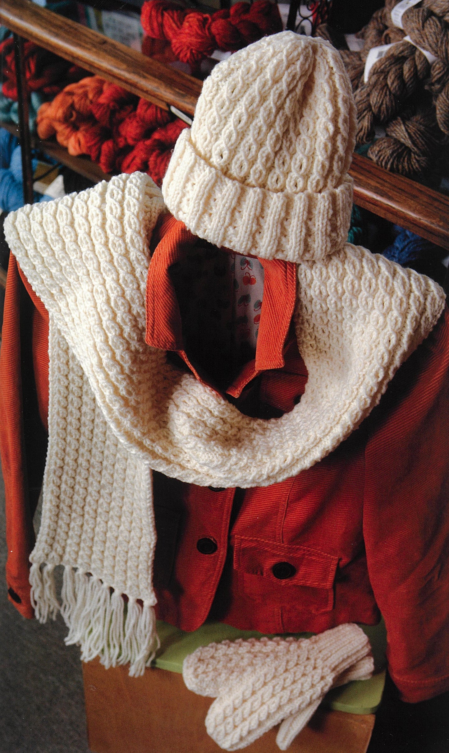 A winter outfit with a red coat and matching cream knit hat, scarf, and mittens is displayed on a chair, with colorful yarn skeins and the Knit Along with Debbie Macomber—Back on Blossom Street book by Leisure Arts in the background.