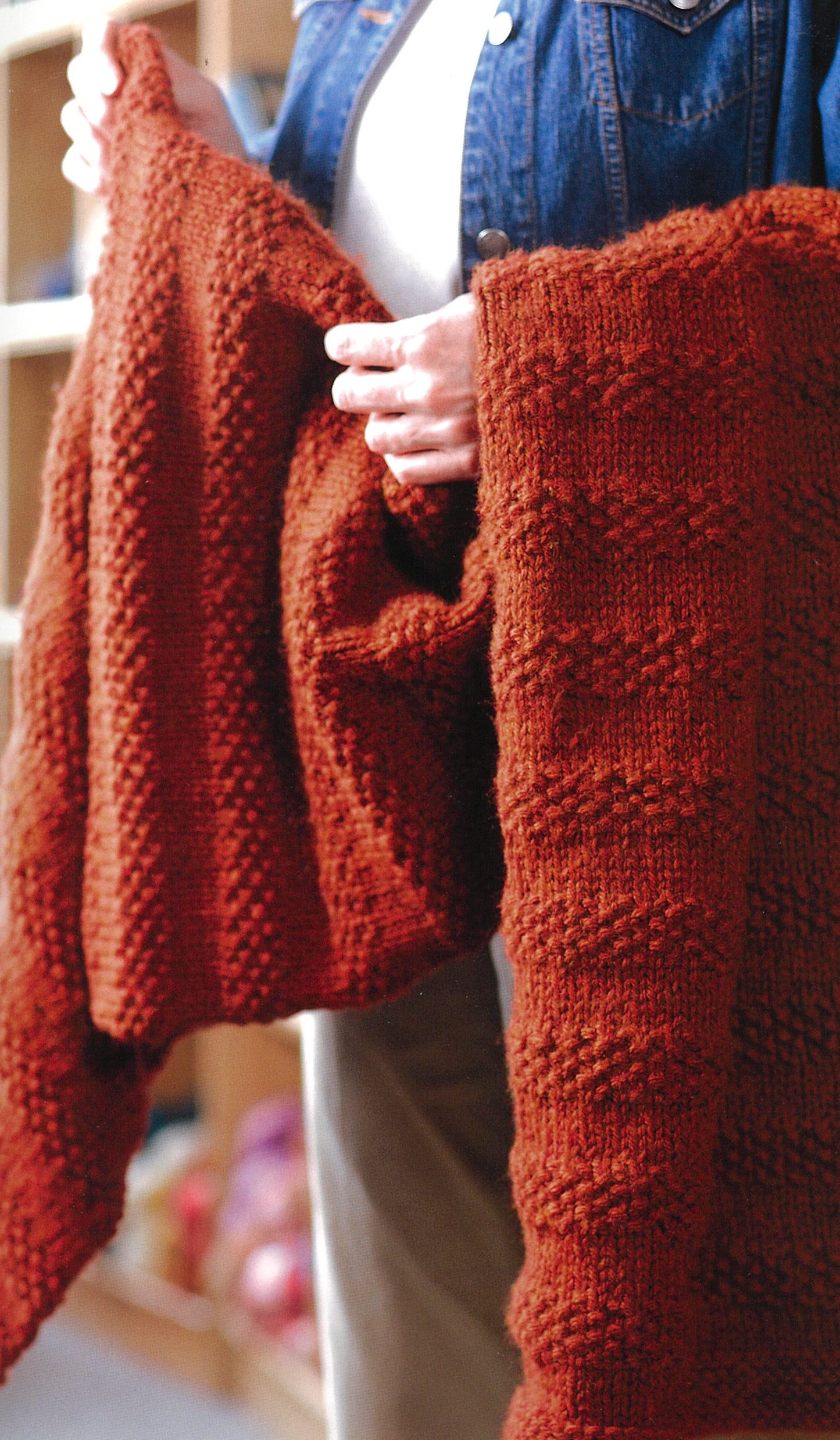 In a cozy indoor setting, a person in a denim jacket displays a rust-red knitted Prayer Shawl, featured in "Knit Along with Debbie Macomber—Back on Blossom Street" by Leisure Arts—the perfect project for any fan.