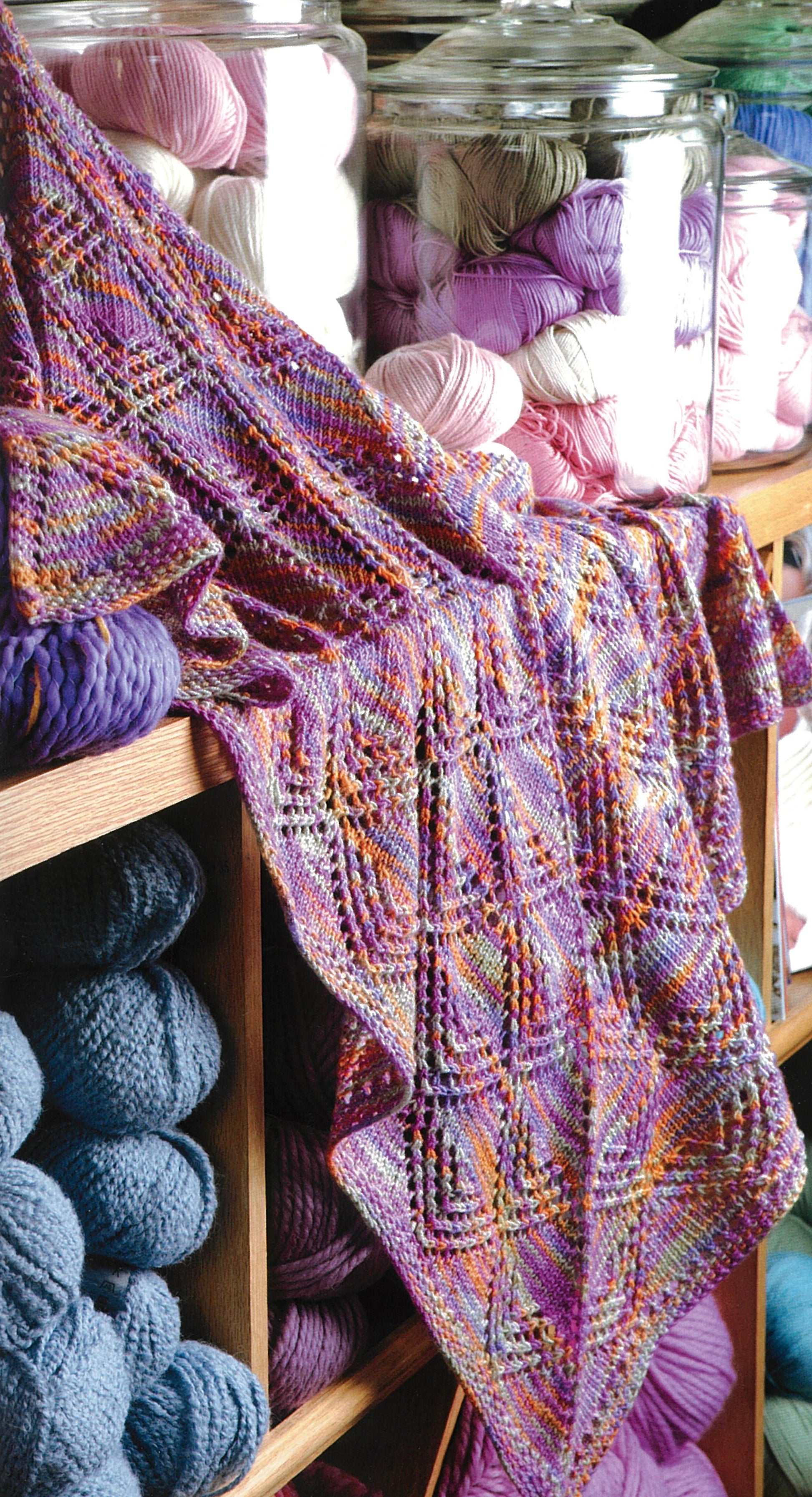 A multicolored lace-knit prayer shawl drapes over a shelf of skeins in blue, pink, white, and purple—ideal for fans of Knit Along with Debbie Macomber—Back on Blossom Street by Leisure Arts.
