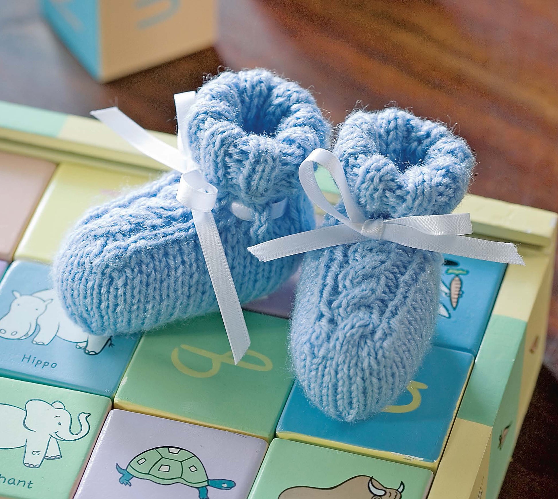 A pair of blue knitted baby booties with white ribbons, made using cable knitting patterns from "I Can't Believe I'm Knitting Cables" by Leisure Arts, are displayed on pastel alphabet blocks featuring animal designs.