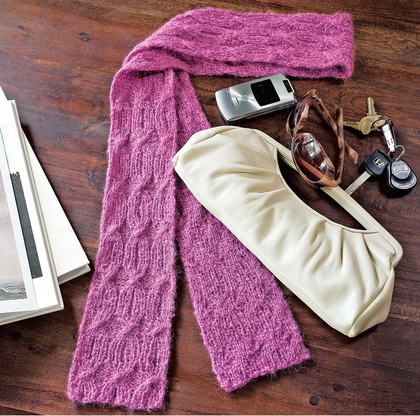 The book "I Can't Believe I'm Knitting Cables" by Leisure Arts is displayed on a wooden surface alongside a pink cable-knit scarf, a white handbag, sunglasses, a flip phone, car keys, and stacked magazines.