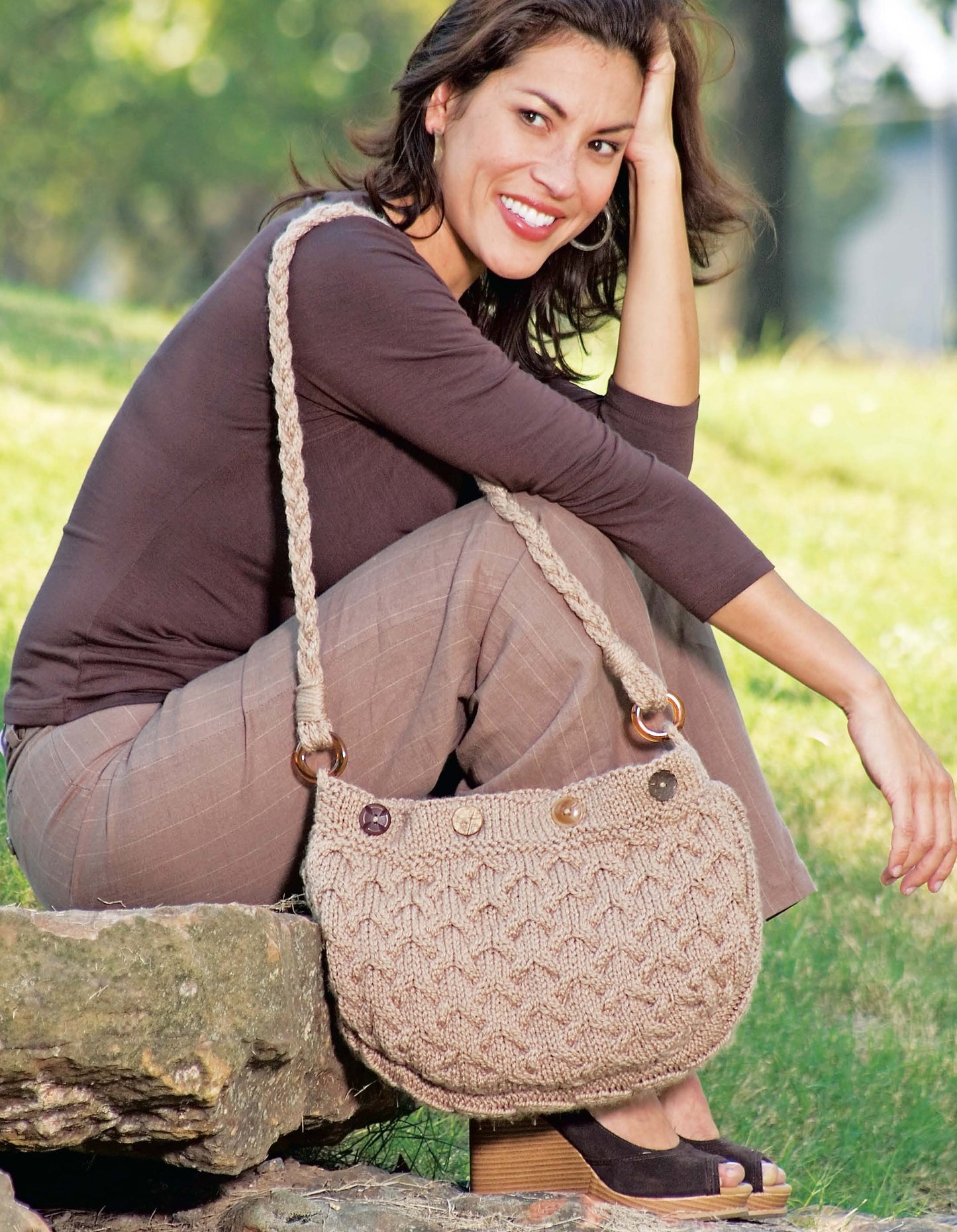 A woman outdoors smiles in a brown outfit, holding a beige shoulder bag with intricate cable patterns made from "I Can't Believe I'm Knitting Cables" by Leisure Arts—perfect for cable knitting enthusiasts.